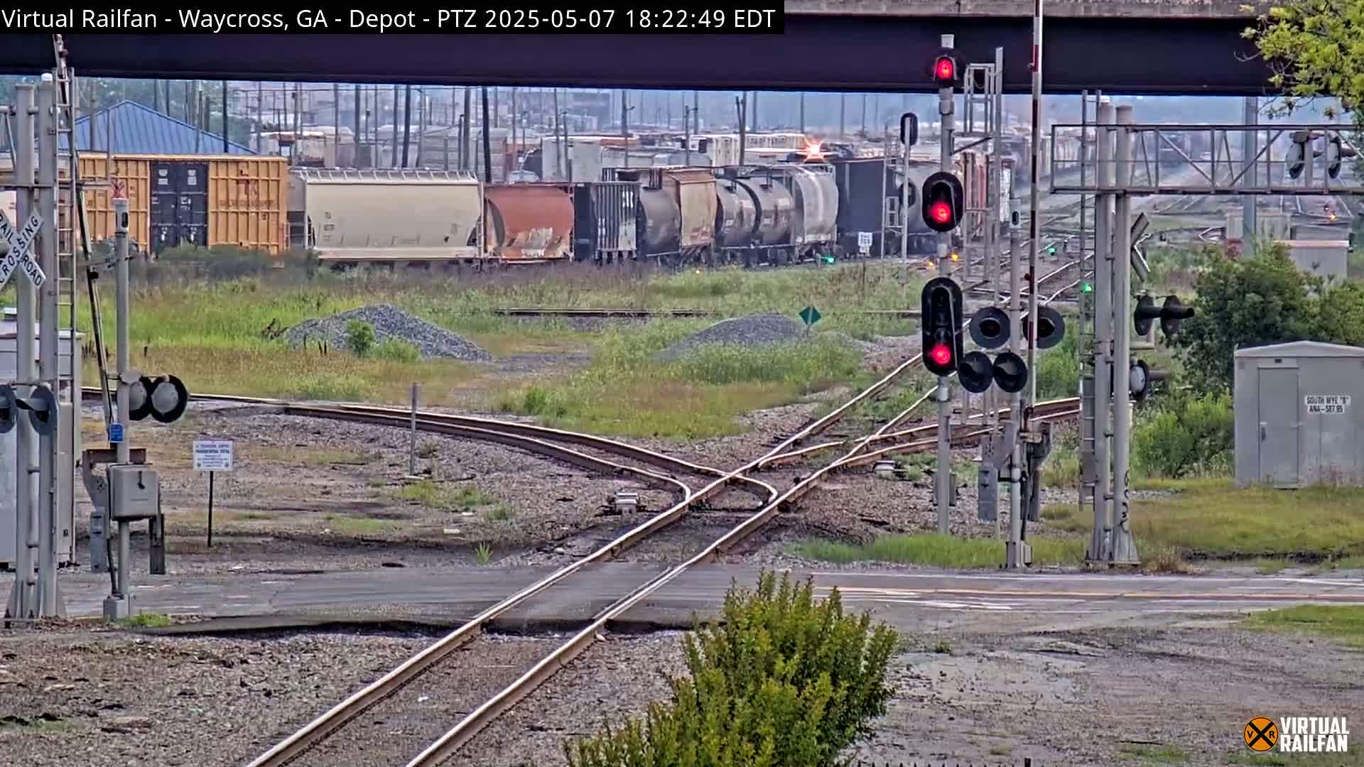A train of boxcars and tank cars sits idle near a railroad crossing with red traffic signals, under an overcast sky.