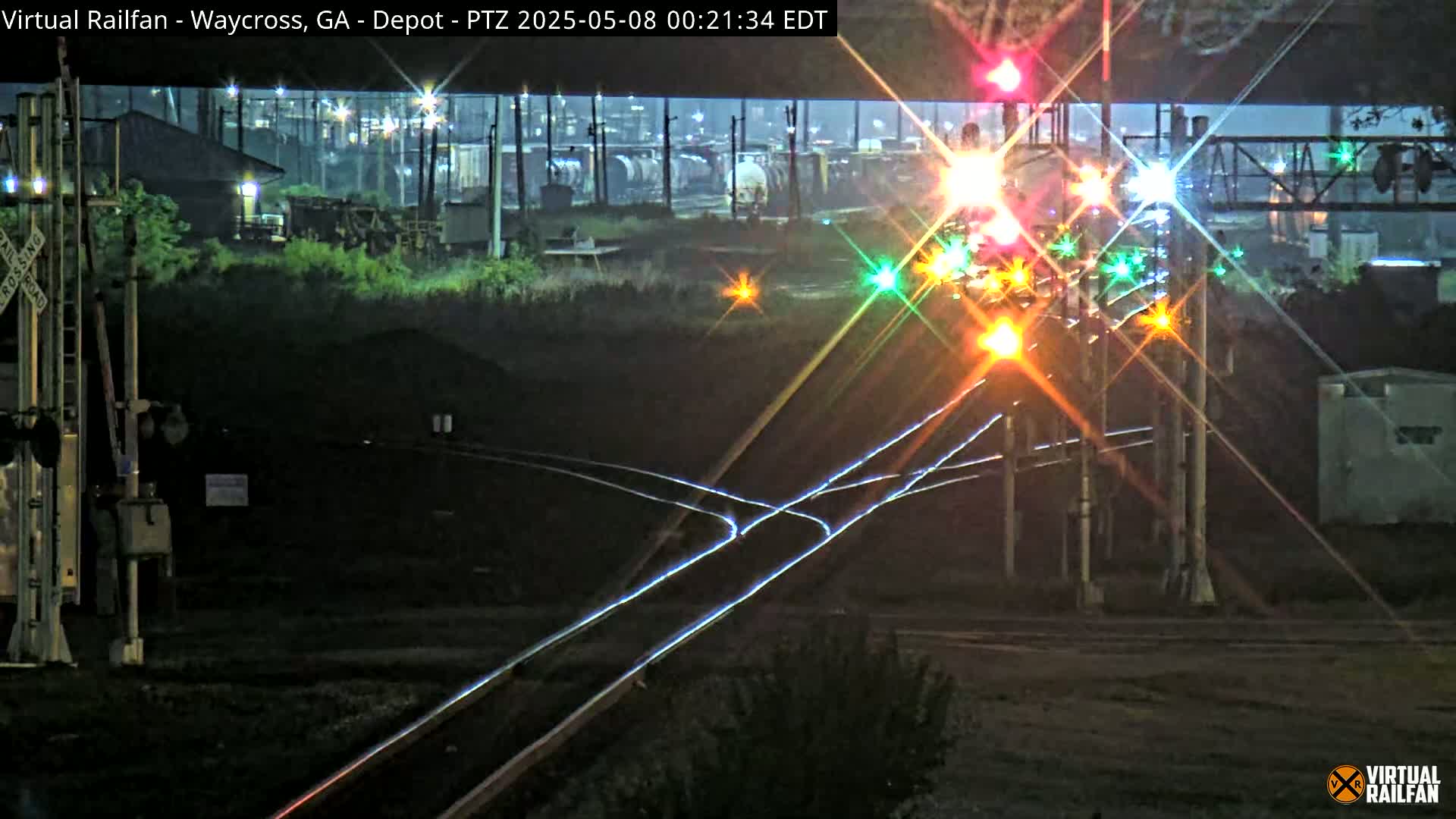 At night, brightly lit railroad crossing signals and tracks intersect near a building and rail cars under a dark sky.