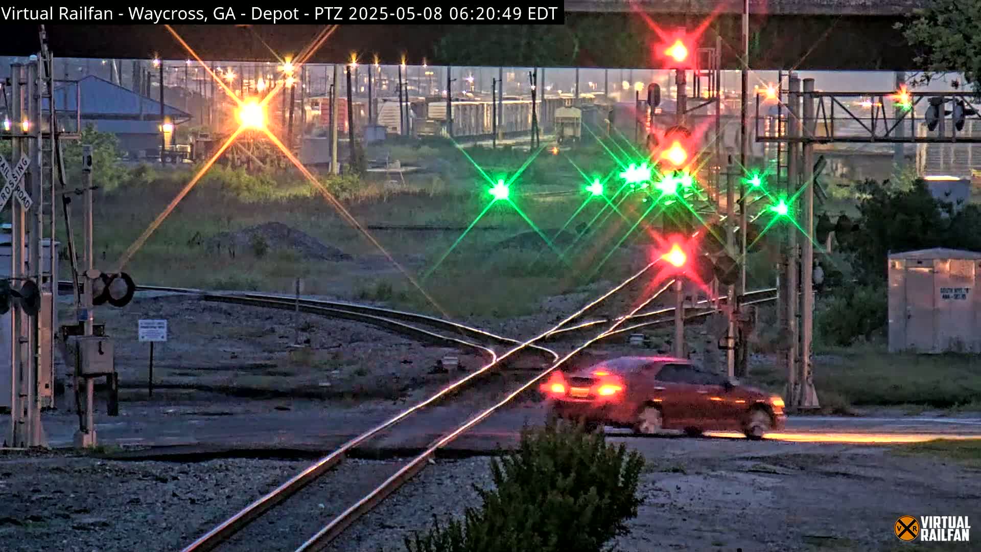 A red car drives across a railroad crossing at twilight, passing under a bridge with active railroad signals displaying red and green lights.