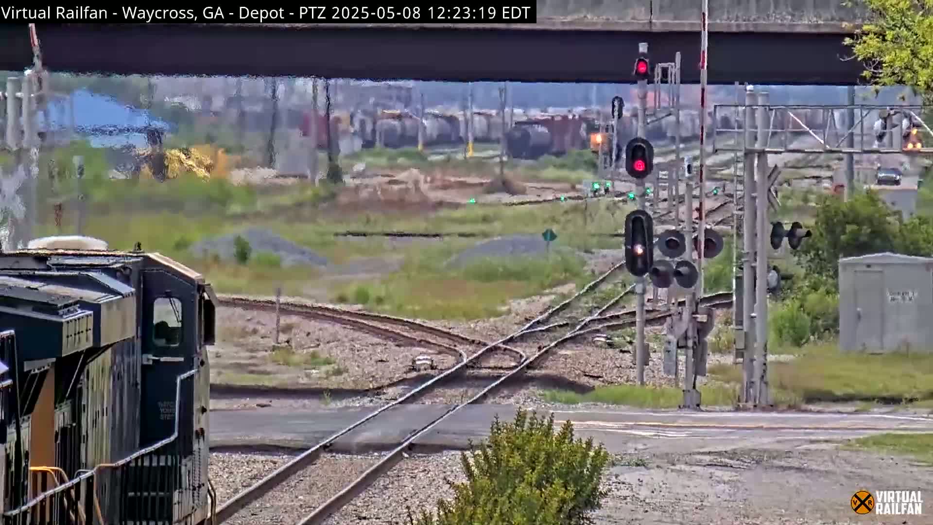 A dark-colored locomotive approaches a railway intersection on a partly sunny day, with multiple sets of train tracks and signal lights visible.