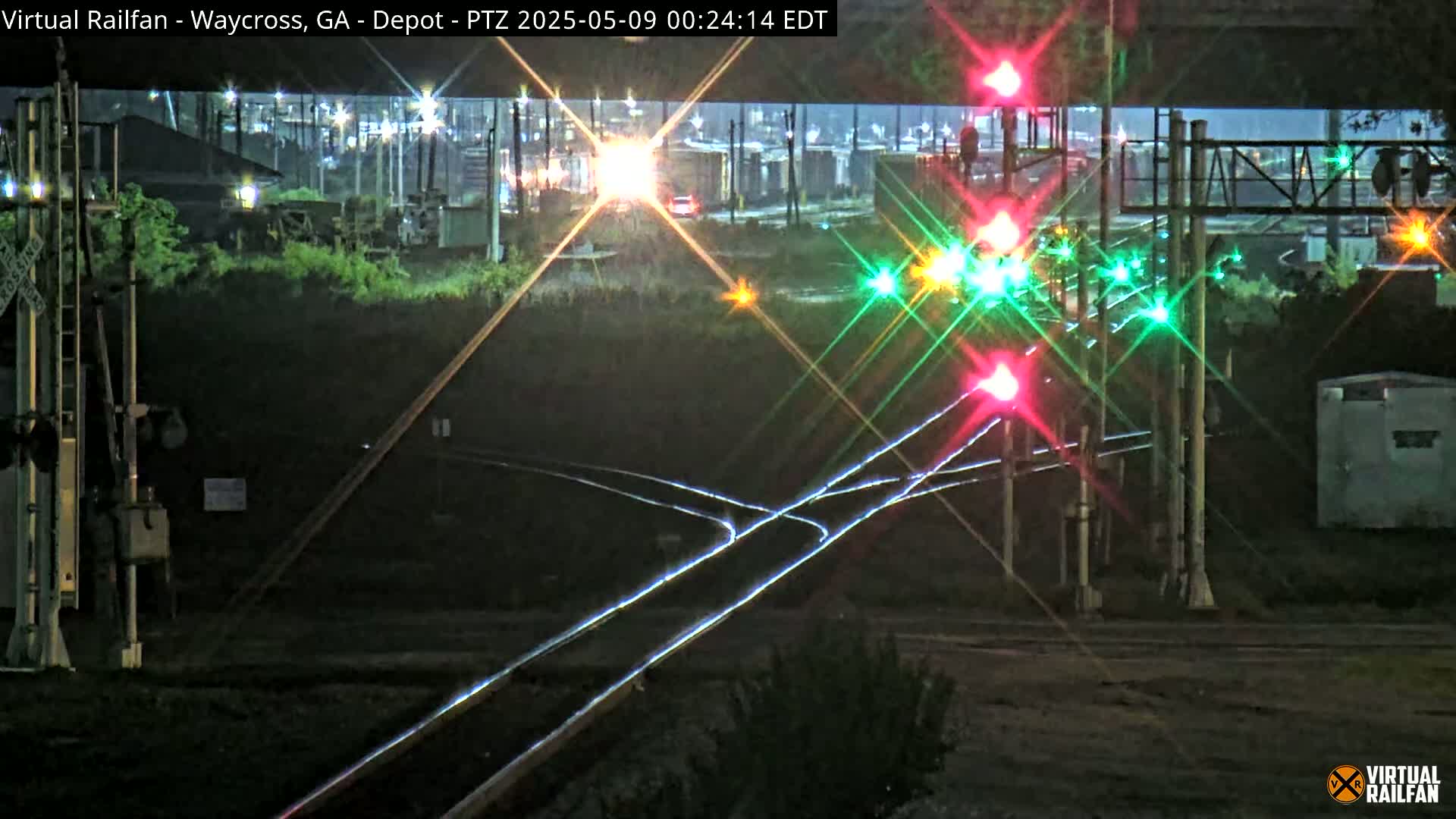 At night, a train passes a railroad crossing with flashing red and green lights under a bridge.