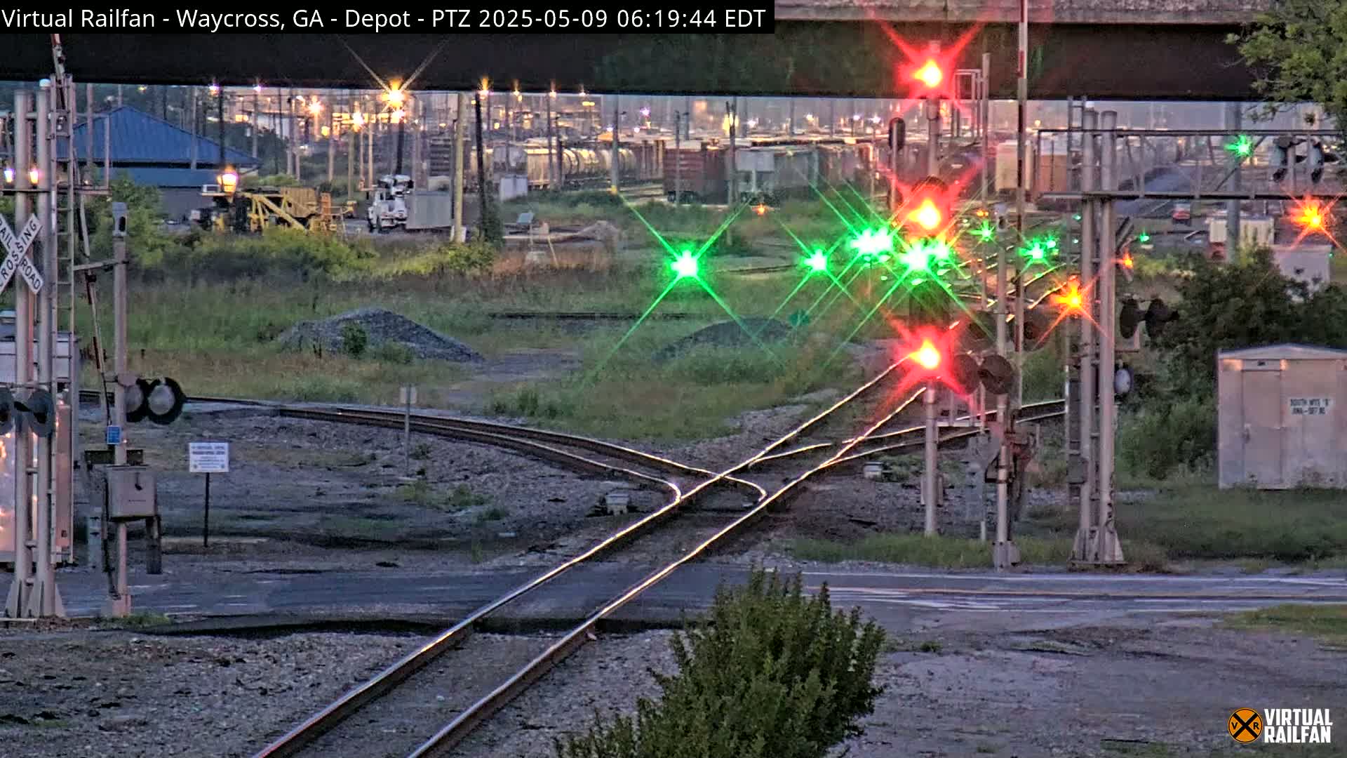 A twilight view of a railroad crossing with multiple train tracks converging, featuring active signal lights and a nearby industrial area.