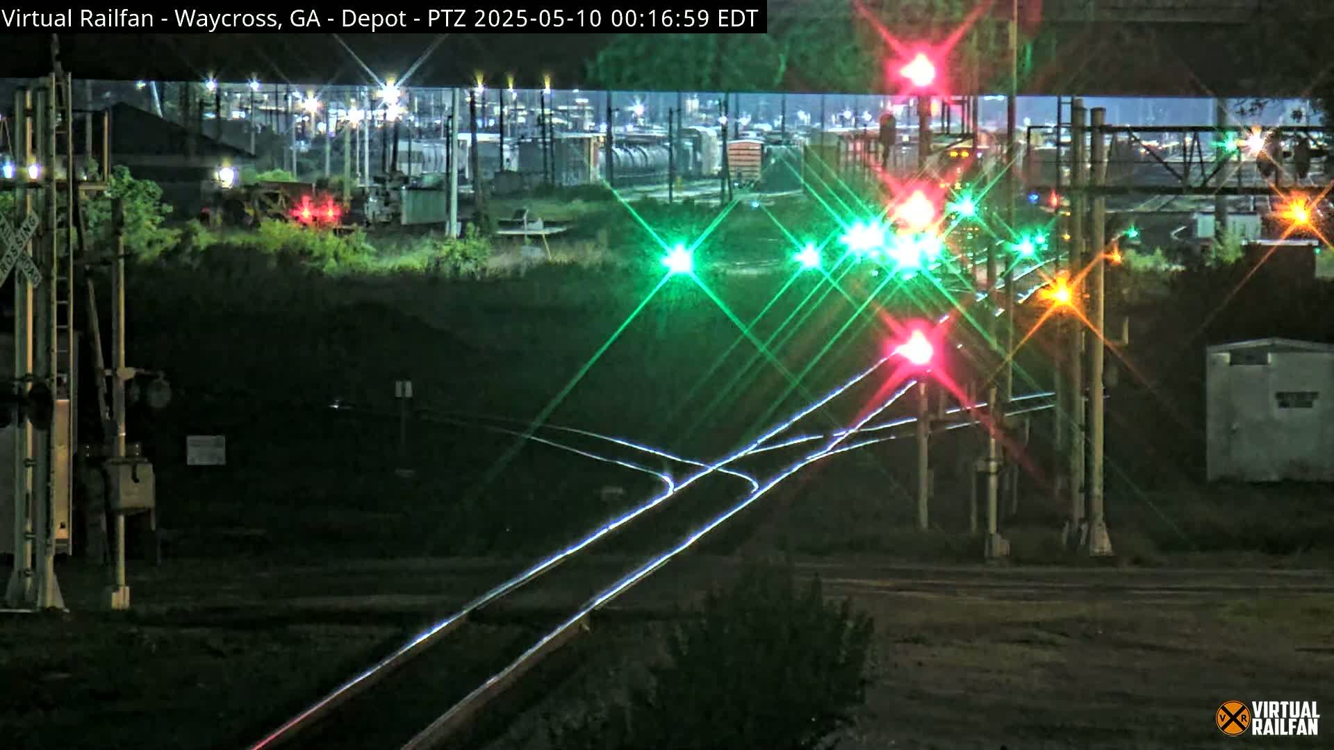 At night, illuminated railroad tracks intersect with a signal displaying red and green lights,  with freight cars visible in the background.