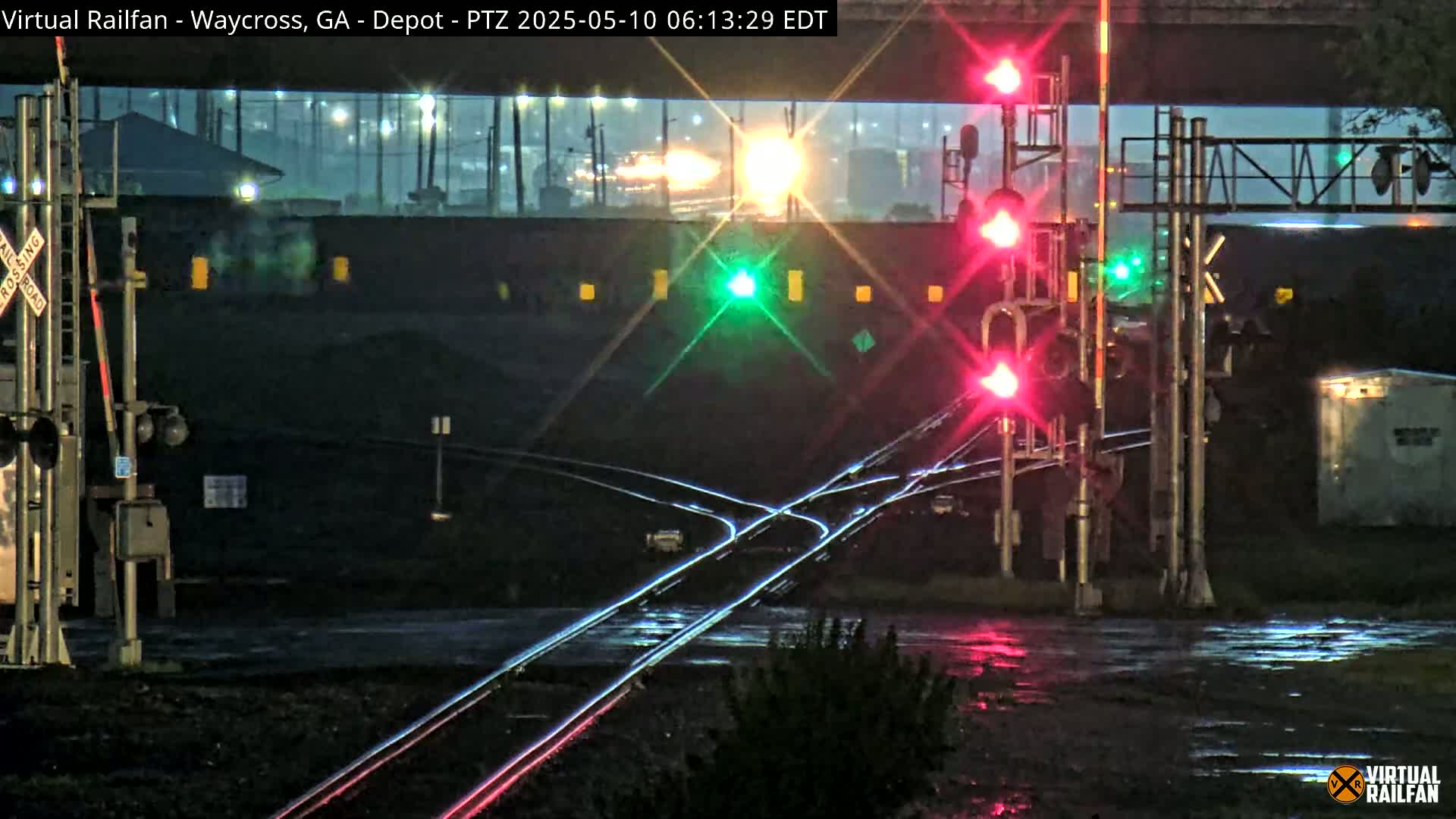 At night in the rain, railroad tracks intersect at a crossing with red and green signal lights and a partially visible train in the background.