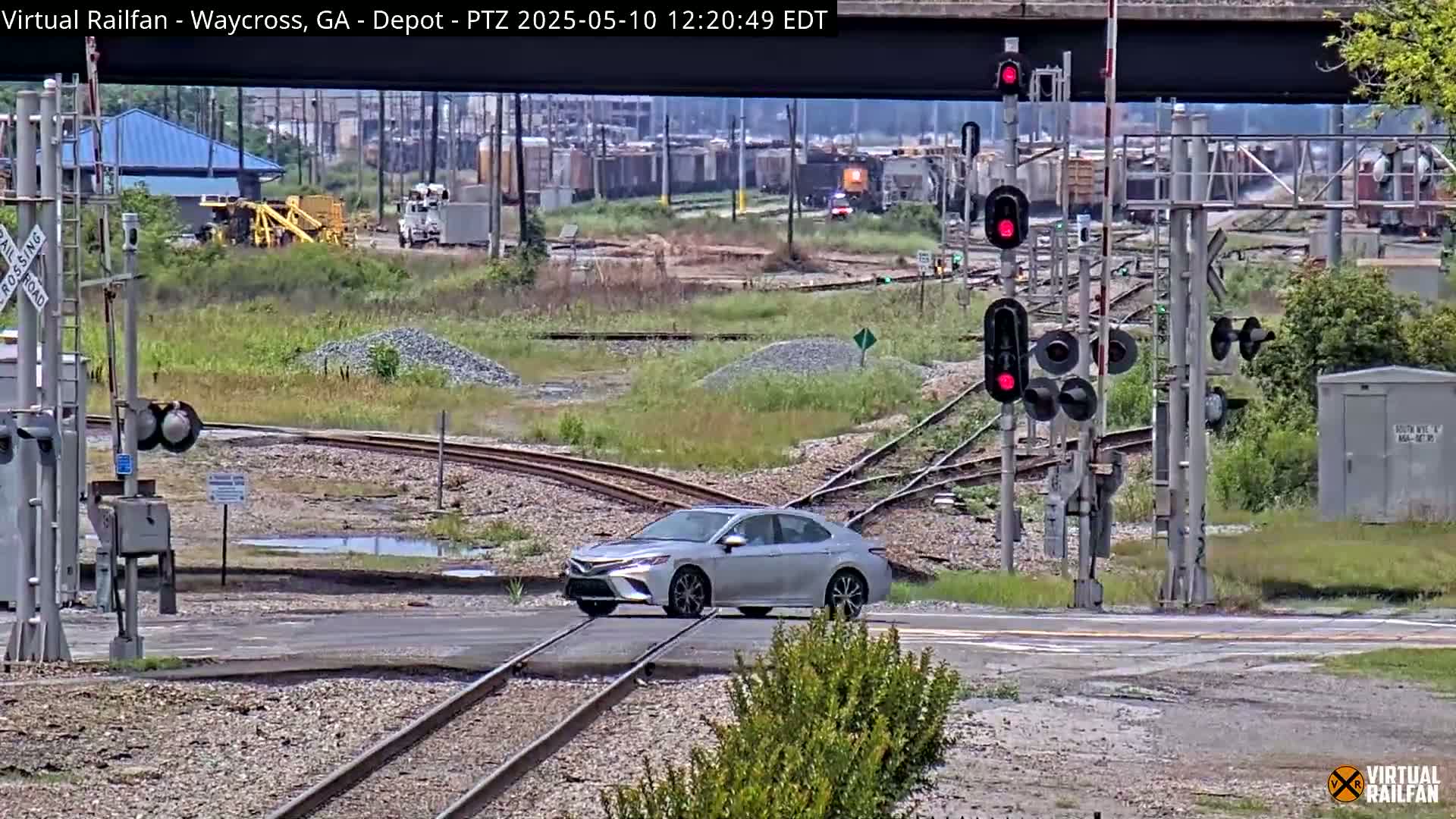 A silver sedan crosses a railroad intersection under a bridge on a partly sunny day.