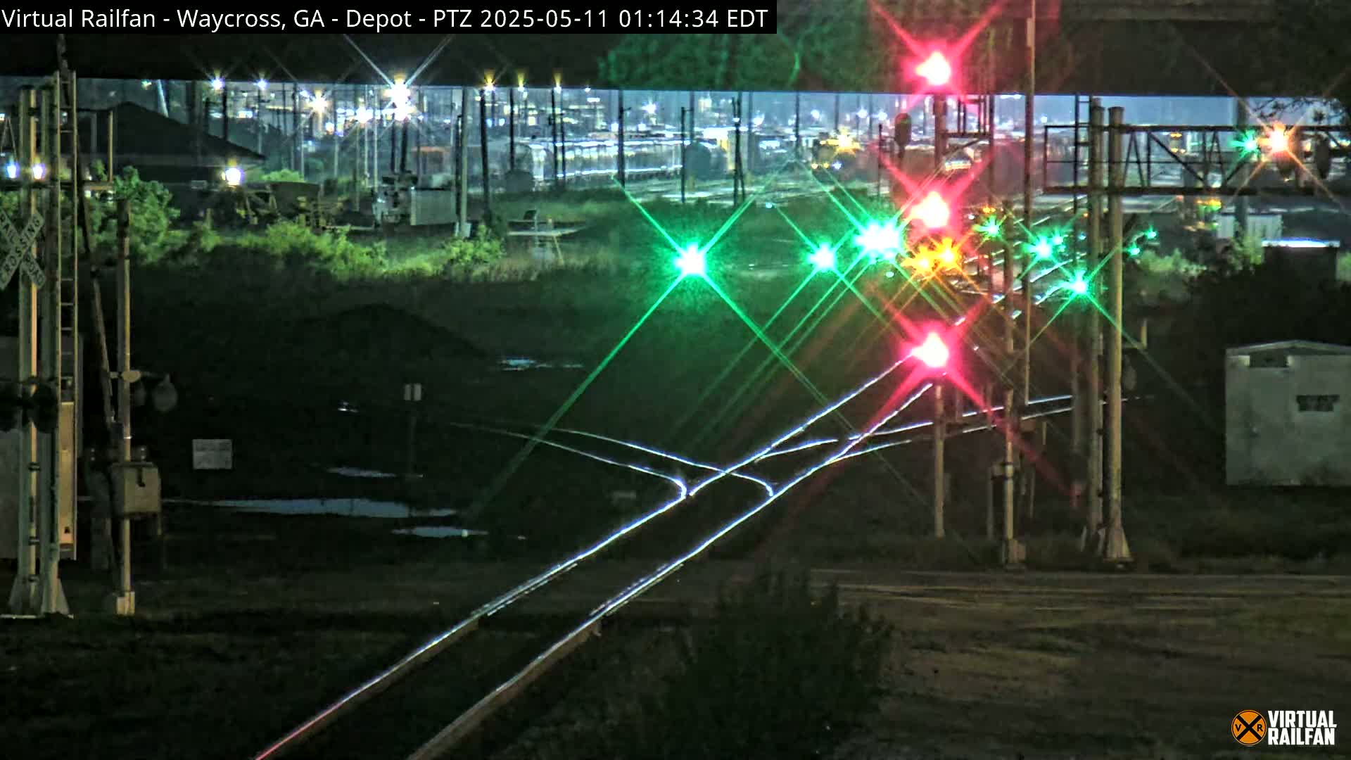 A nighttime view of a railroad crossing with its signals illuminated, showing a train yard in the background.