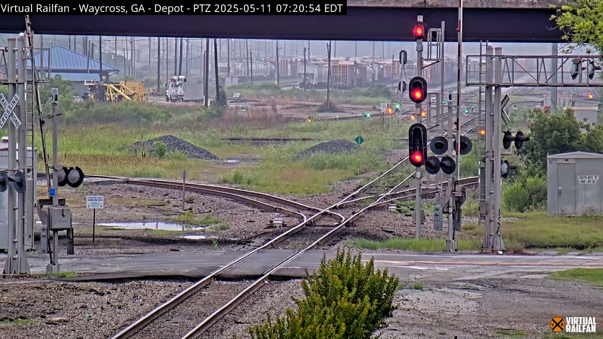 A train yard with multiple sets of railroad tracks intersecting, some railcars in the distance, and several active railroad signals under an overcast sky.