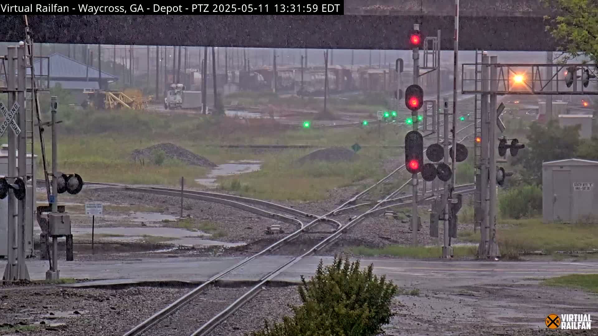 A rainy scene shows a railroad crossing with several sets of tracks converging, traffic signals, and a building in the background.
