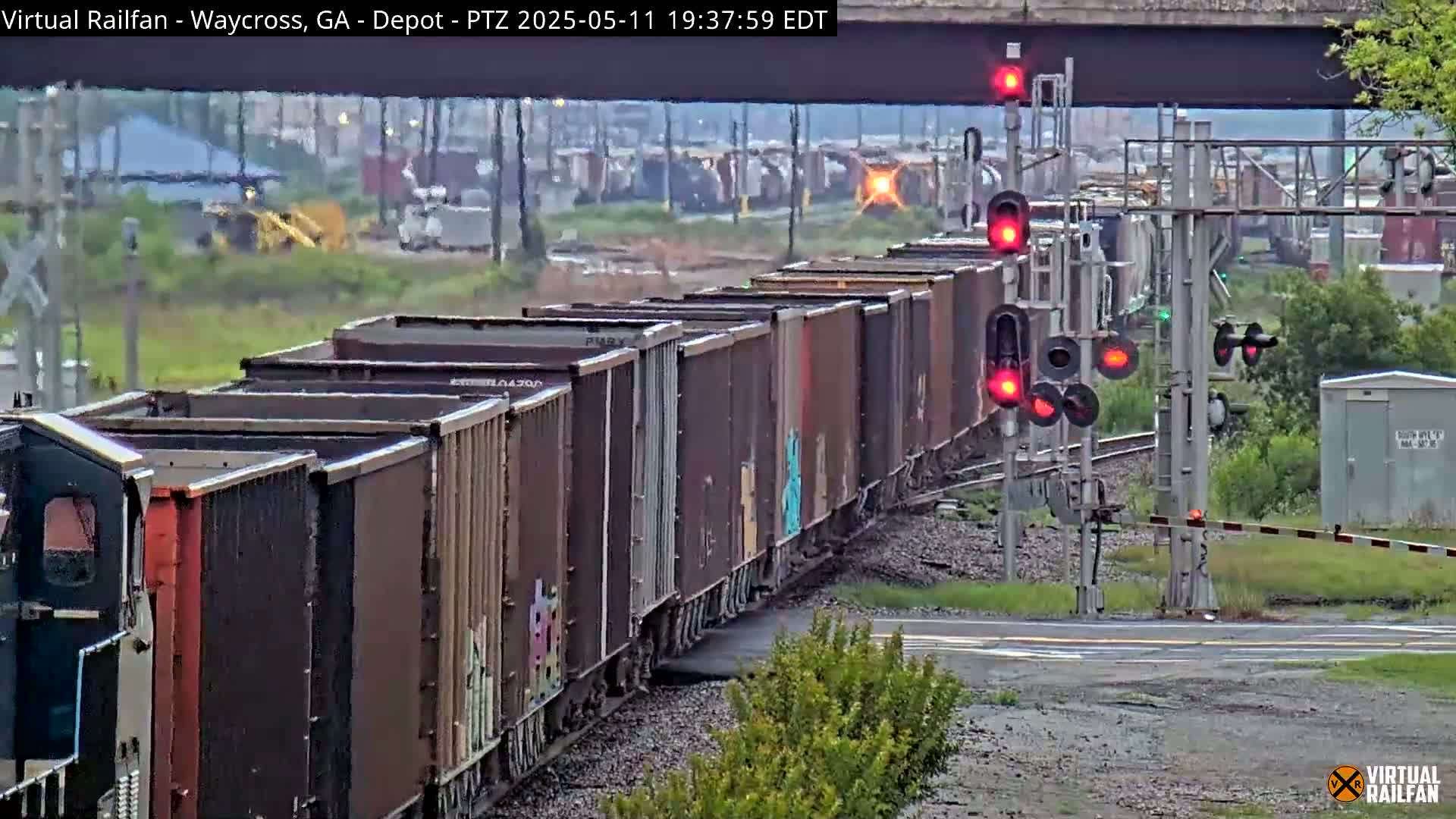 A long freight train with many dark brown boxcars passes a railway crossing under an overcast sky.