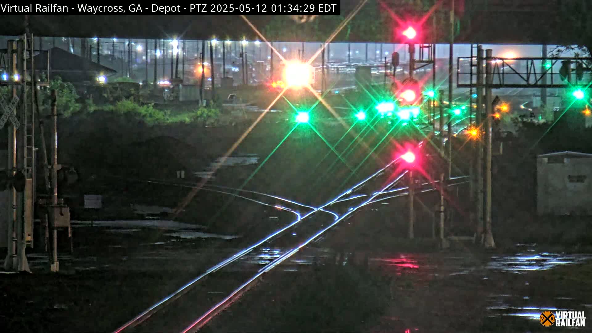 A nighttime view of a rain-slicked railway intersection with several illuminated traffic signals and converging train tracks.