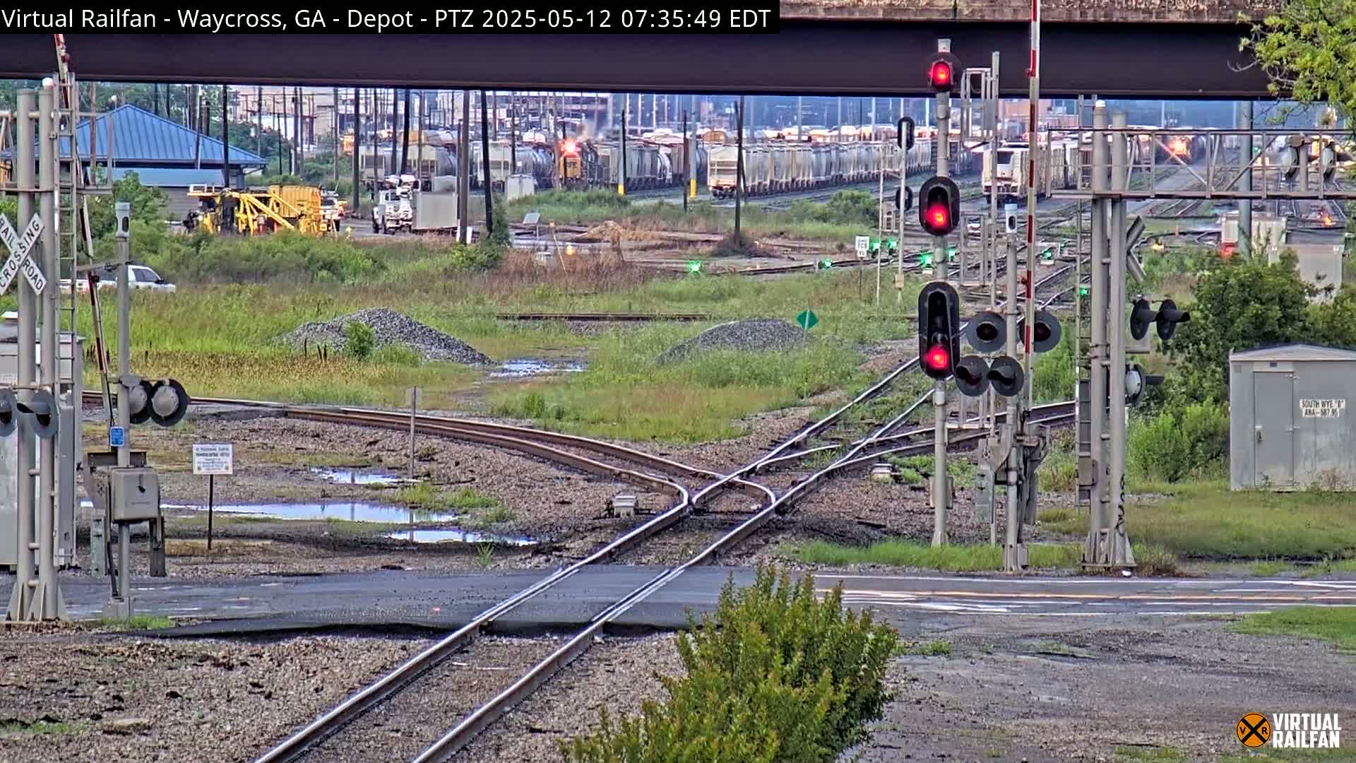 A railway intersection with several train cars in the distance, under an overcast sky, shows puddles of water near the tracks.