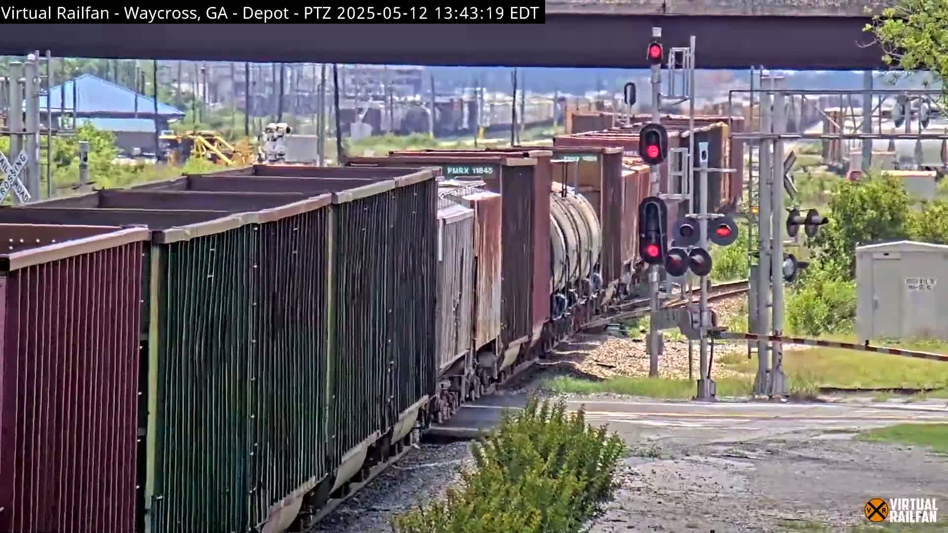 A long freight train with various cargo cars passes a railroad crossing under a bridge on a partly sunny day.