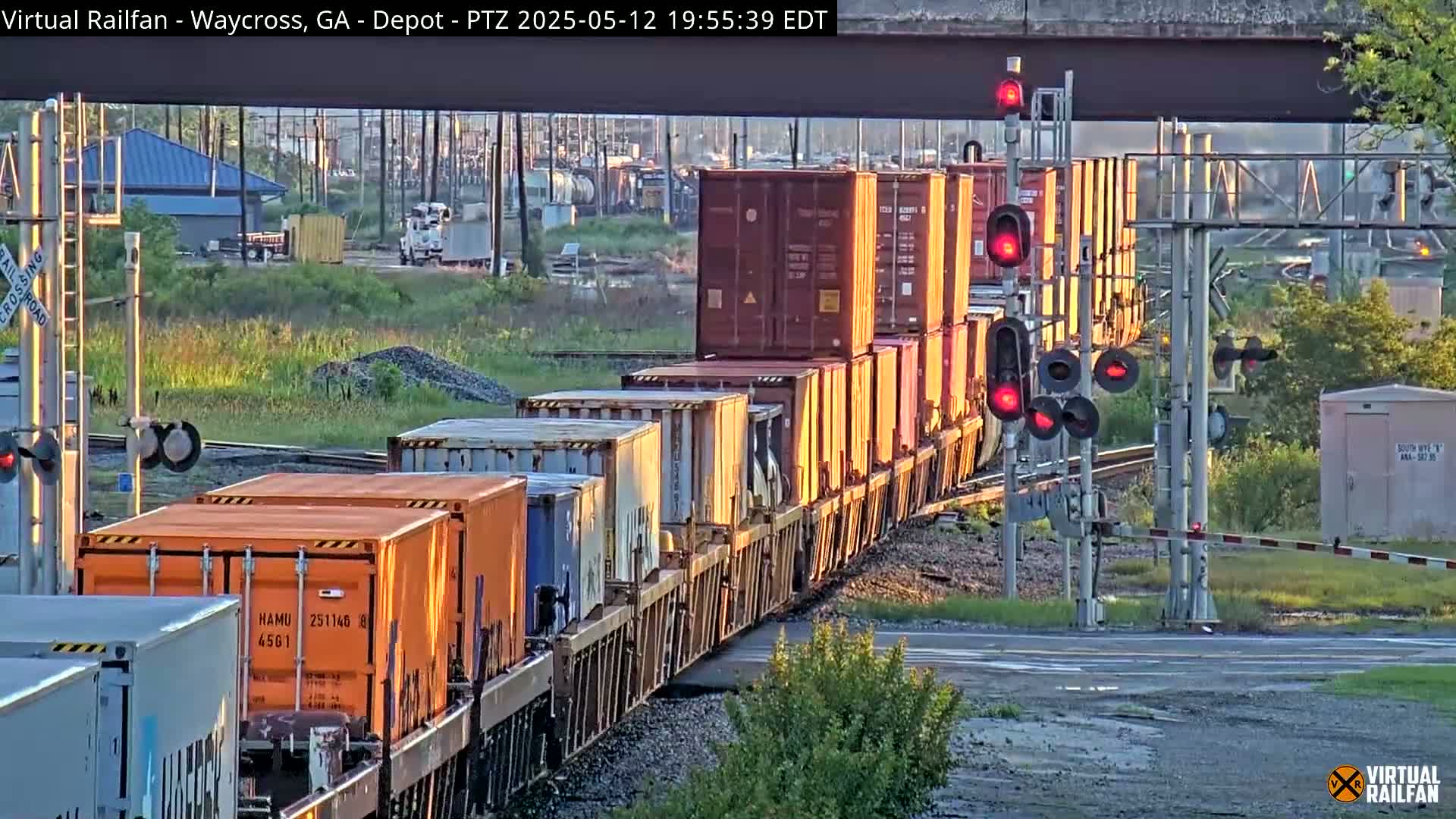 A long freight train passes a railroad crossing under a bridge at sunset.