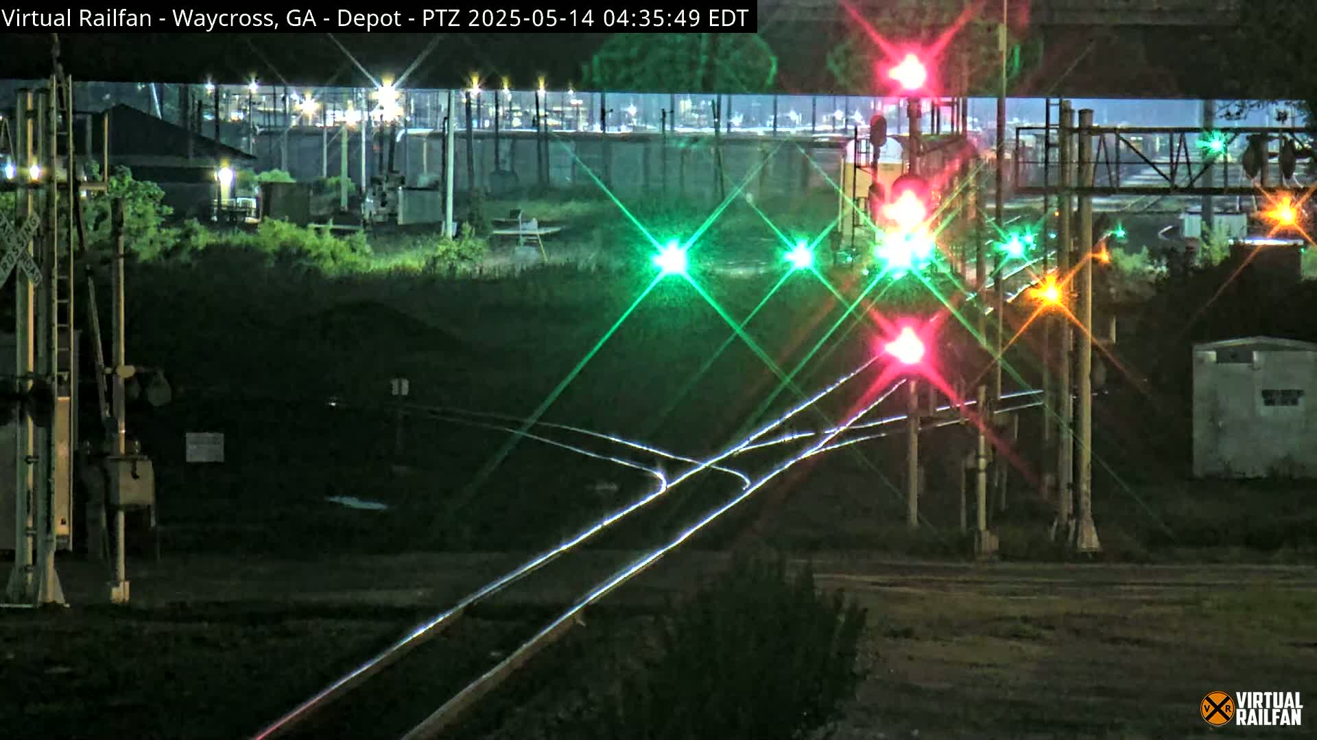 At night, under a dark sky, a nighttime view shows a railroad crossing with several illuminated signal lights and train tracks.