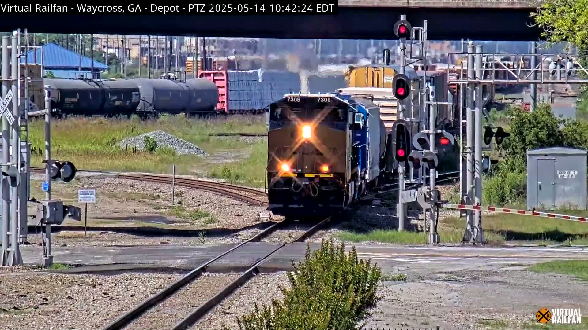 A blue and brown locomotive leads a freight train across a railroad crossing on a sunny day.