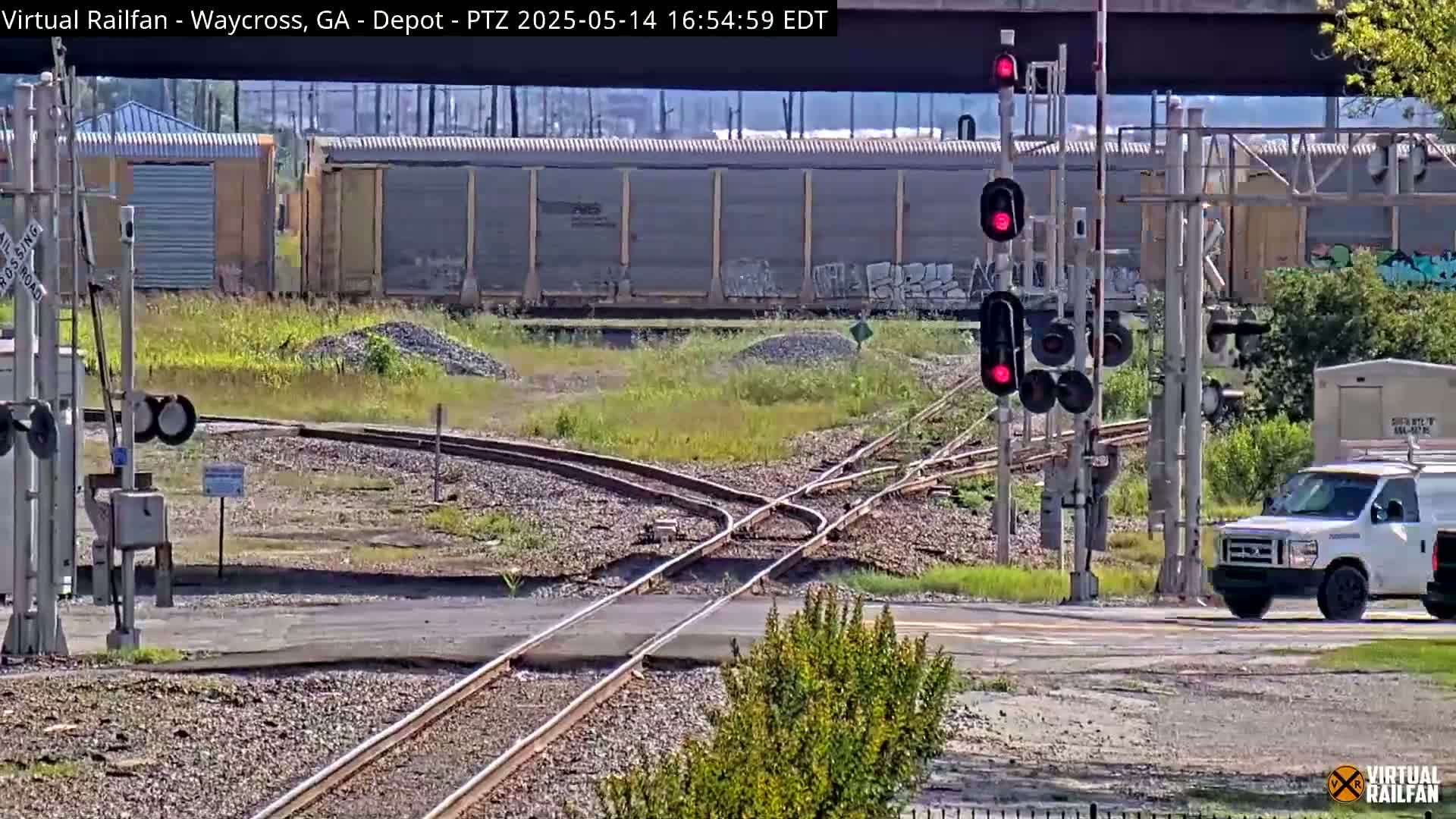A sunny day at a railroad crossing shows a white van stopped at a signal, with train tracks diverging, and several railcars in the background.