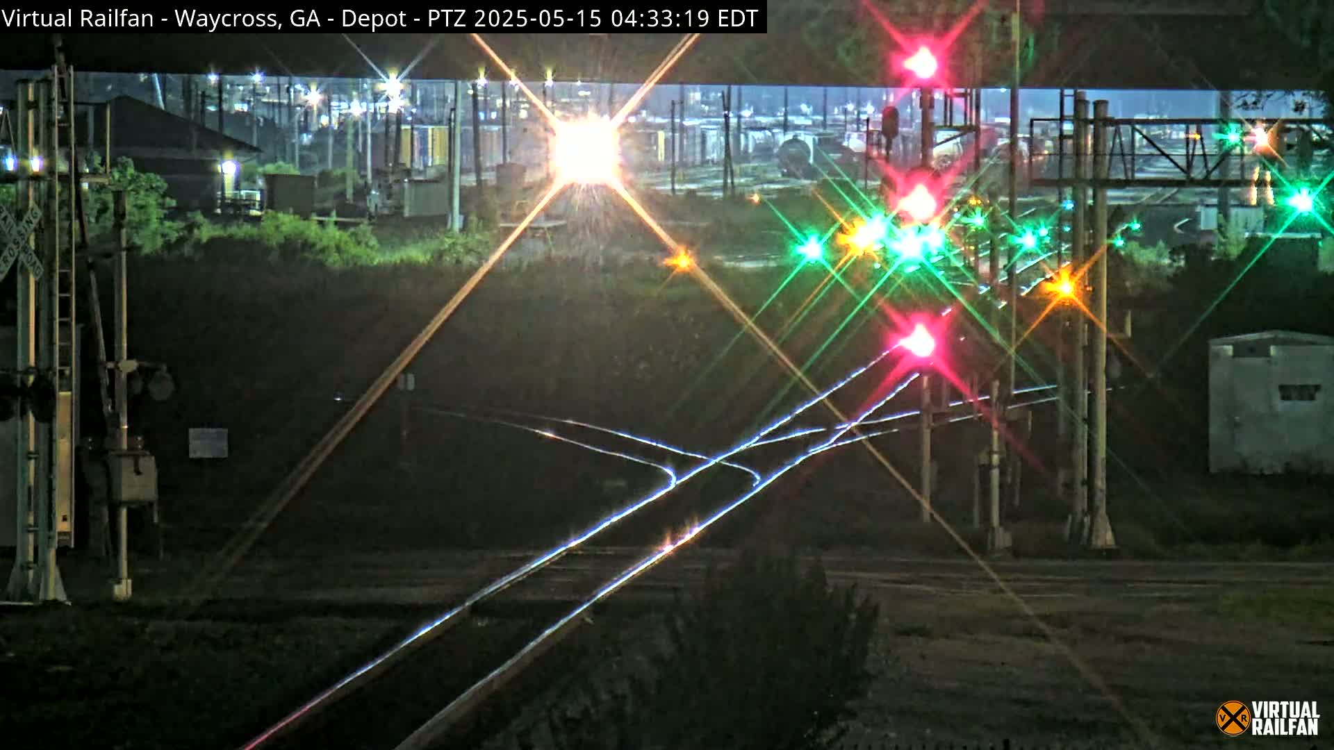 At night, a train crossing with illuminated signals and railroad tracks is shown, with some distant lights and buildings visible in the background.