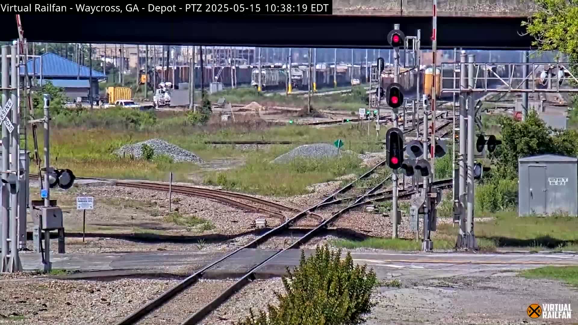 A sunny day at a railroad crossing shows a complex network of train tracks intersecting with a road, various railroad signals, and some industrial buildings in the distance.