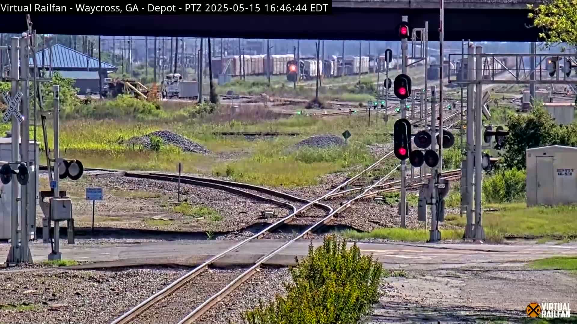A railway crossing with several sets of red traffic signals, train tracks branching off in multiple directions, and a freight train in the distance under a partly sunny sky.