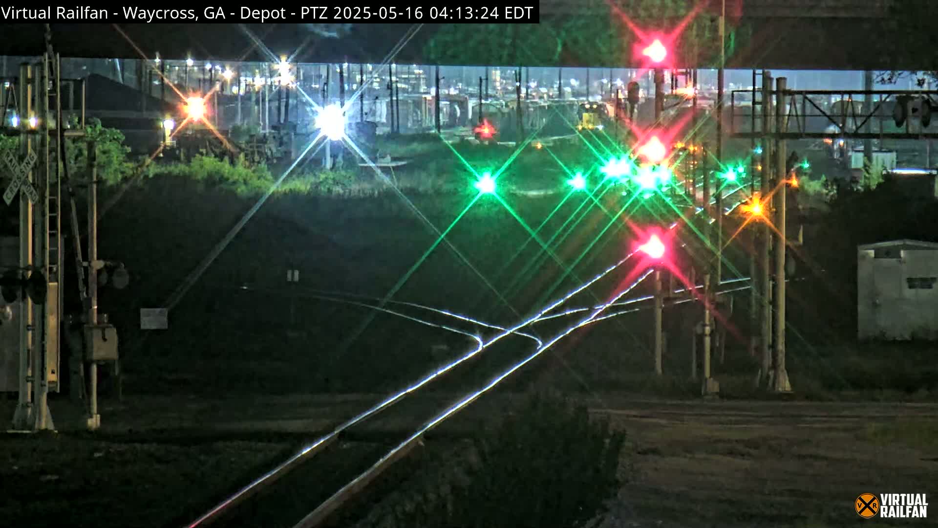 A nighttime view of a railroad crossing with illuminated signals, train tracks, and a distant cityscape under a dark sky.