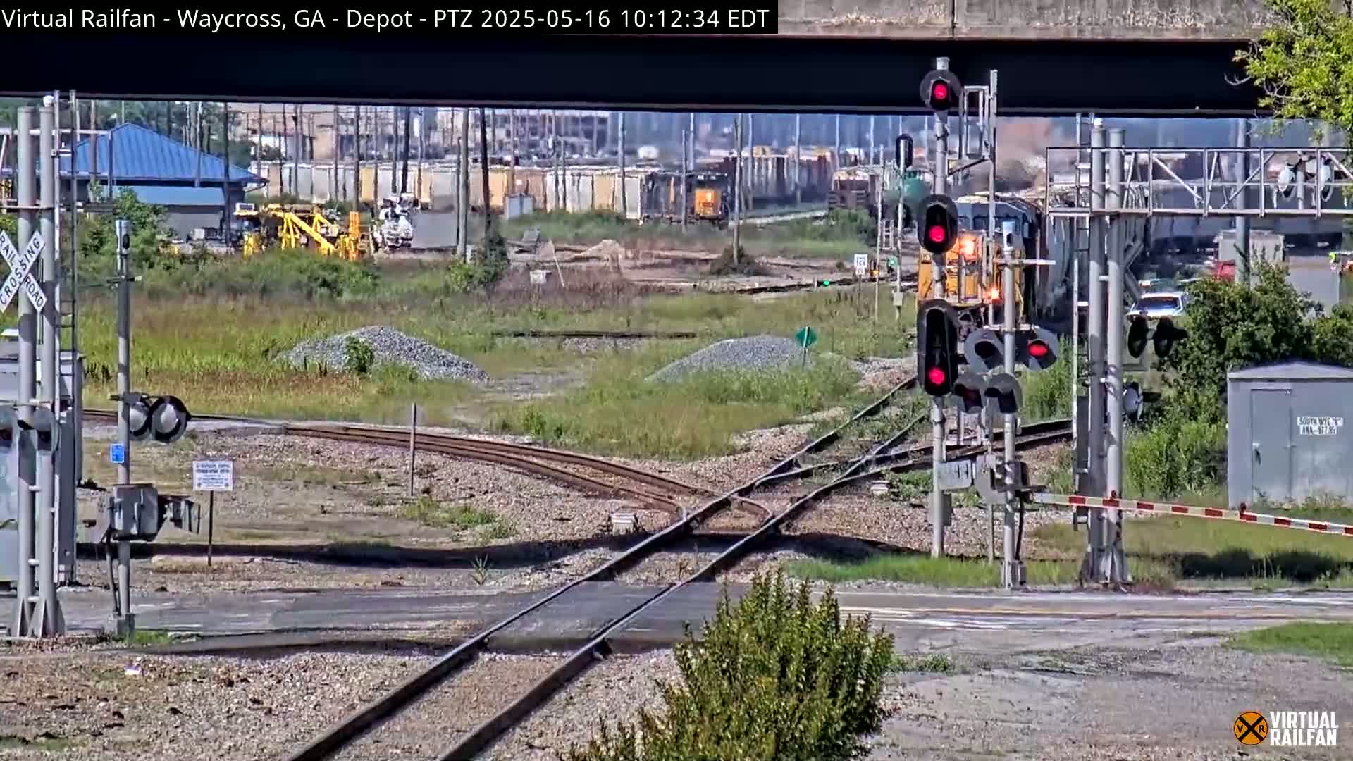 A yellow locomotive leads a train past a railroad crossing with red signal lights activated, under a partly sunny sky.