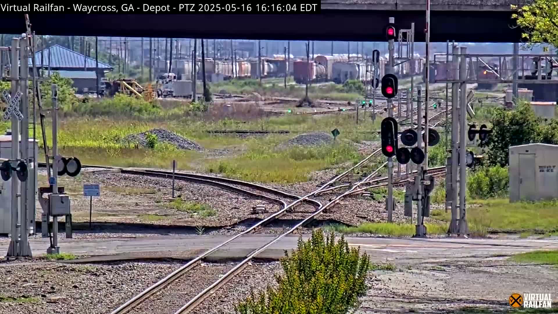 A sunny outdoor view shows a railroad crossing with several sets of tracks, some rail cars in the distance, and red traffic signals indicating a stop.
