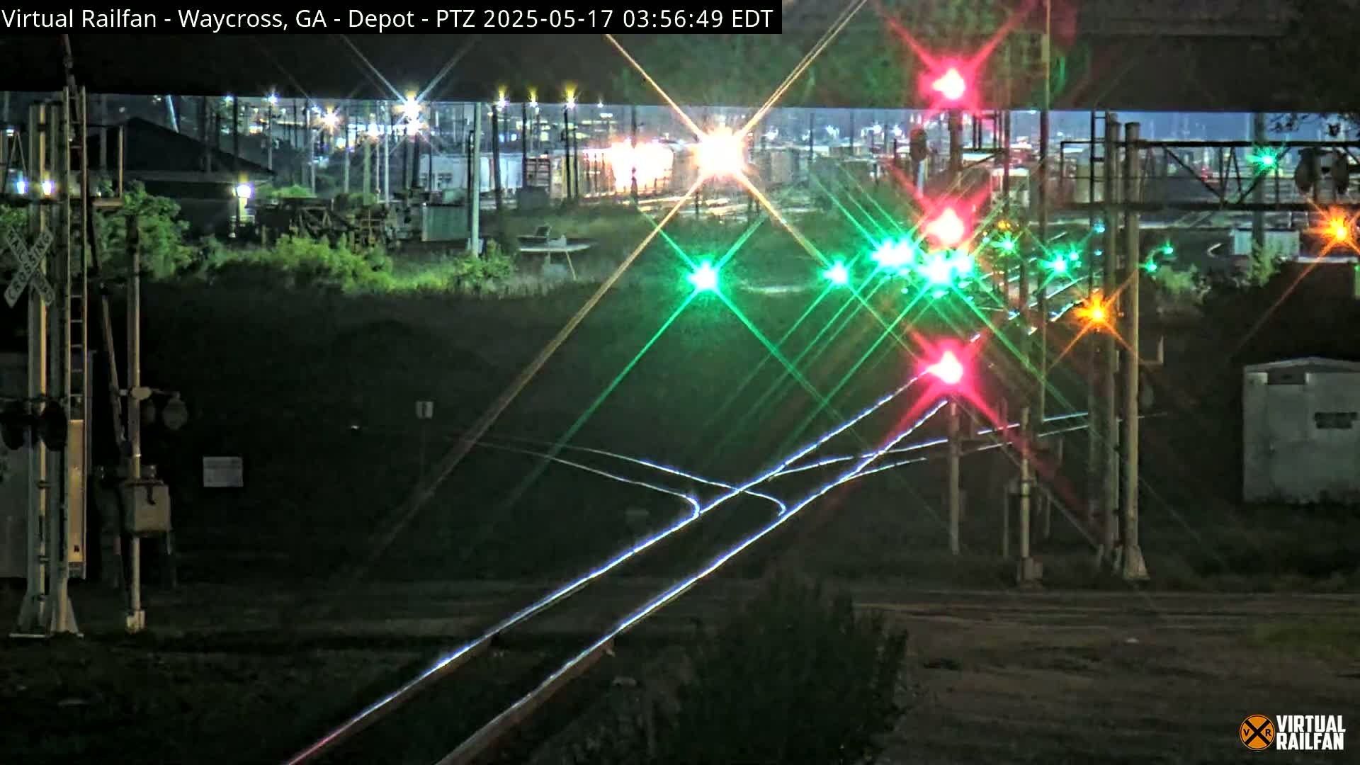 At night, a train crossing with several brightly lit traffic signals shows green and red lights reflecting on the railroad tracks.