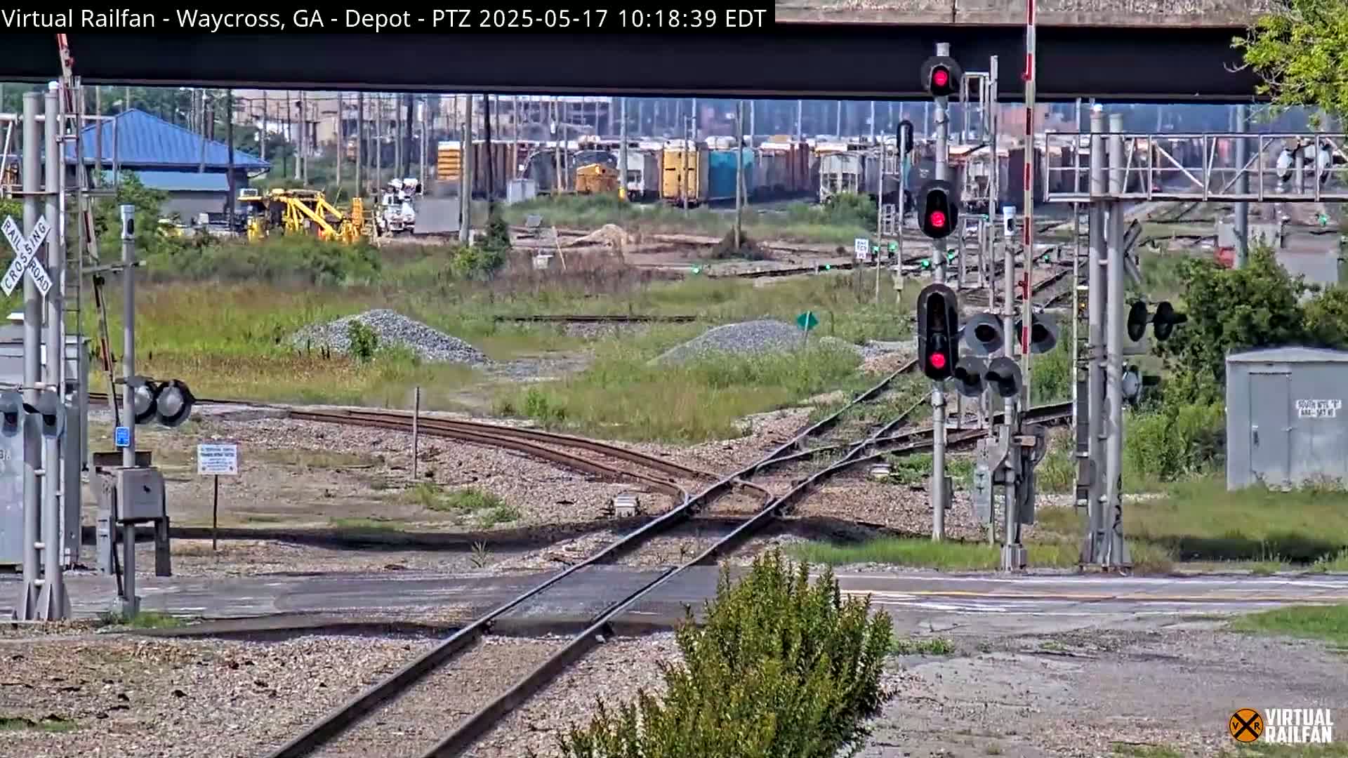 A railroad crossing with several sets of tracks, railway signals displaying red lights, and a yard with parked railcars, all under a partly sunny sky.