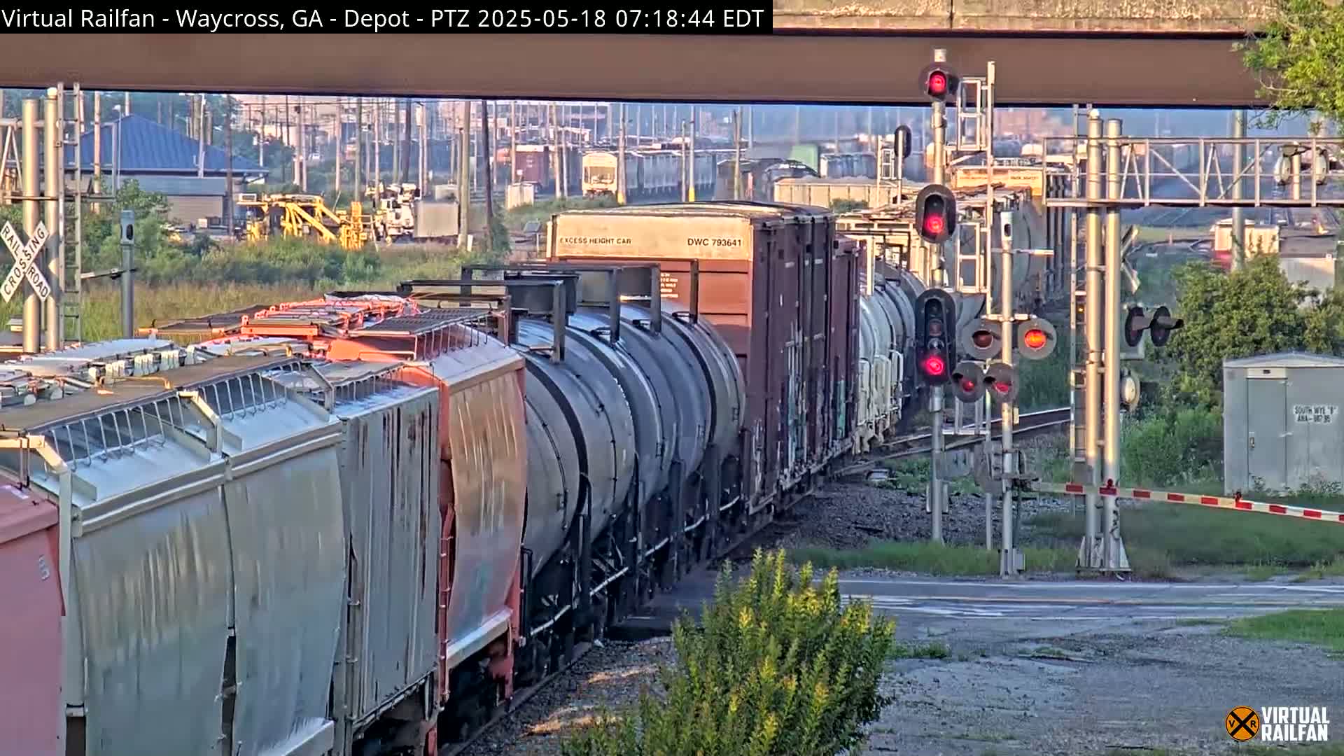 A long freight train with several tank cars and covered hopper cars passes a railway crossing under a bridge in bright, sunny conditions.