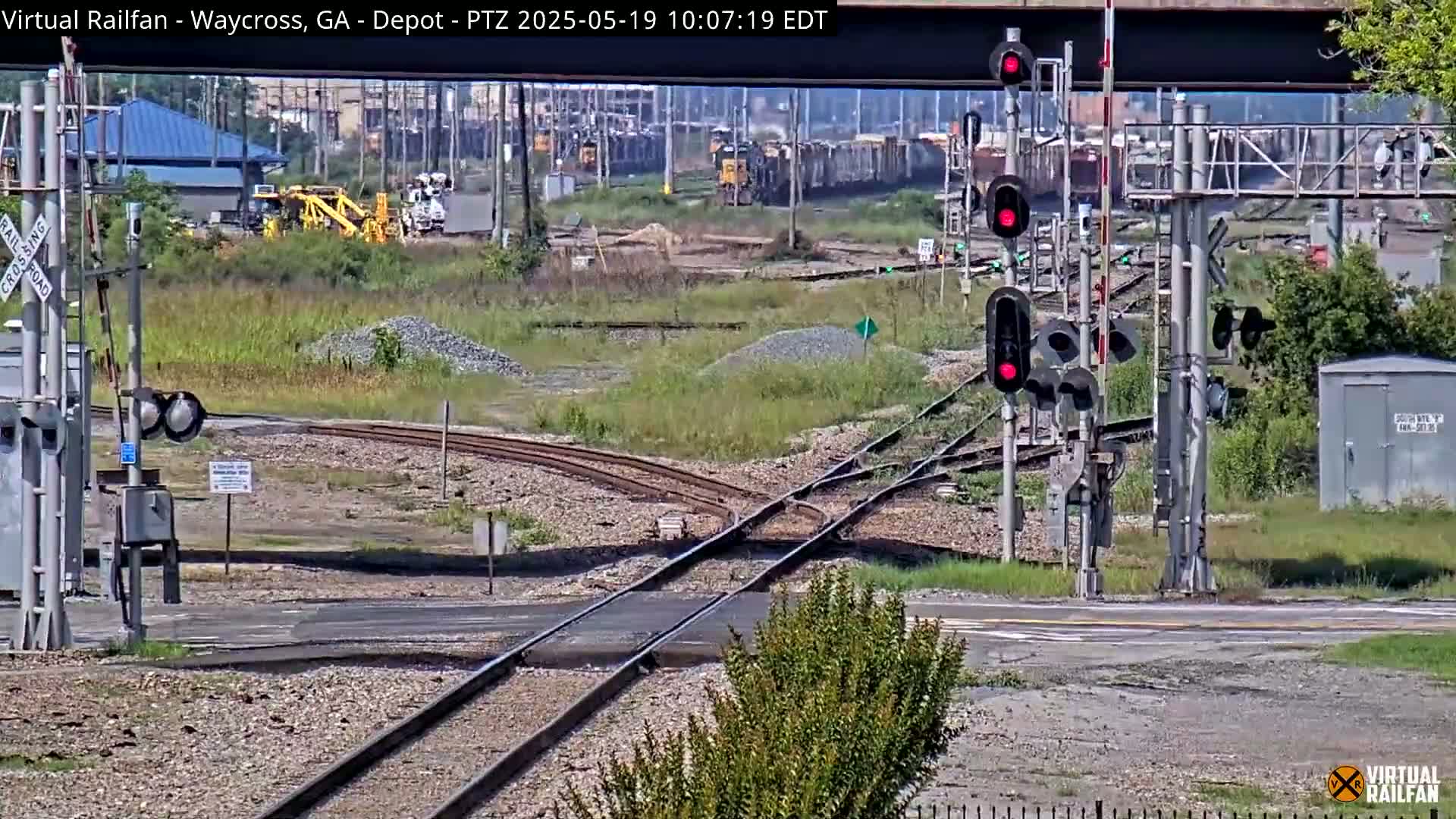 A sunny day shows a railway crossing with multiple train tracks, signals displaying red, and a train in the distance, surrounded by some light vegetation and gravel.