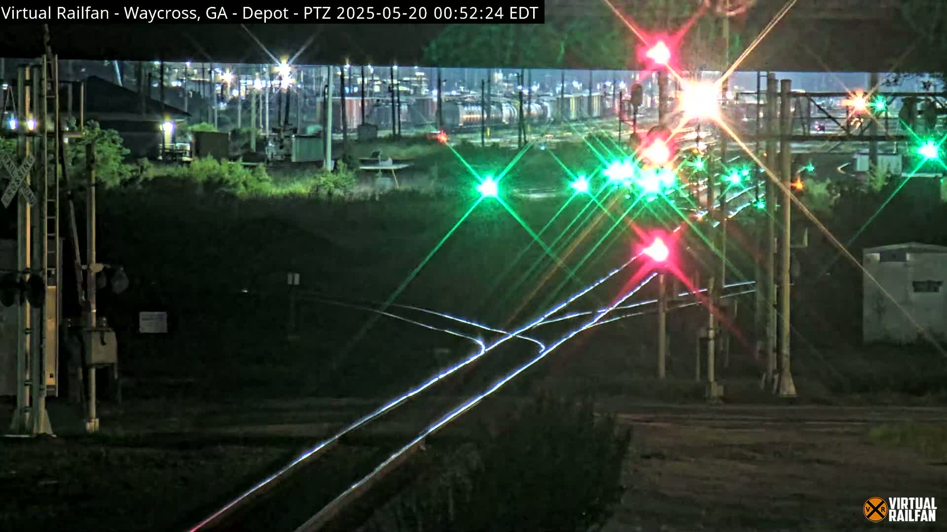 At night, railroad tracks intersect at a depot with multiple flashing red and green signal lights illuminating the scene.