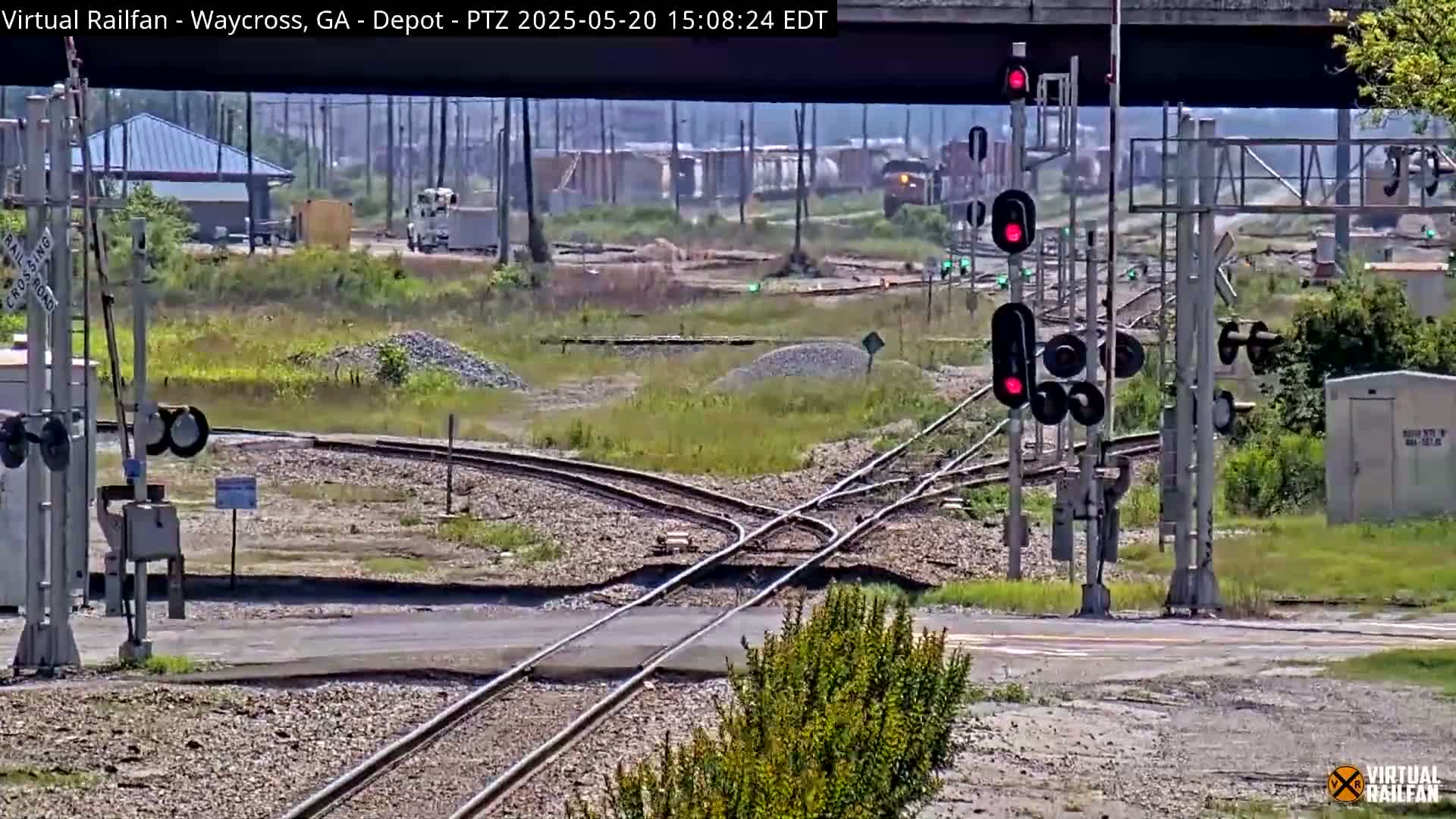 A railroad crossing with several sets of train tracks intersecting, featuring railroad signals and a small building, is visible under a partly cloudy sky.