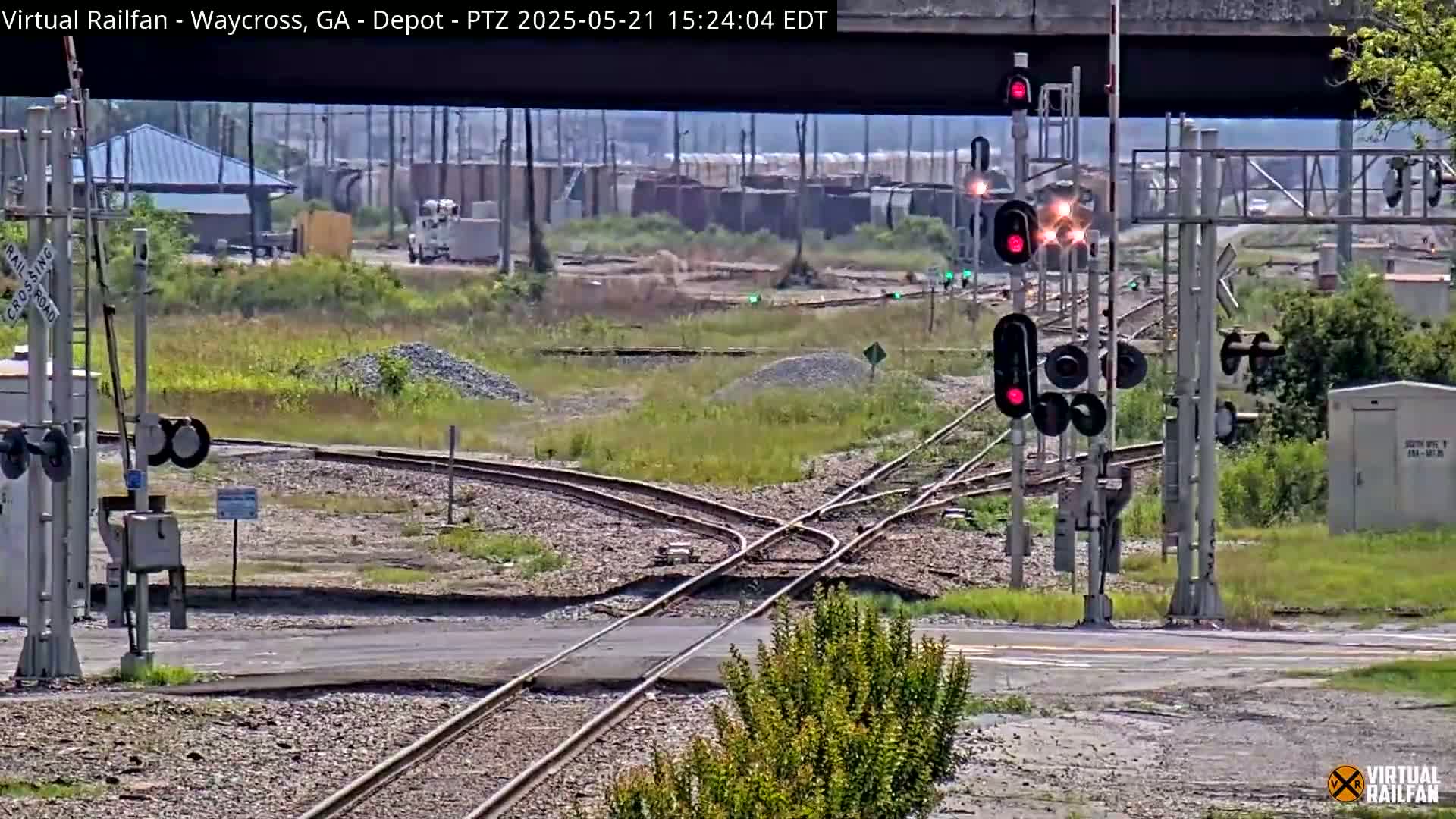 A sunny day at a railroad crossing shows a complex intersection of train tracks with red signal lights, a road crossing the tracks, and some industrial buildings in the background.