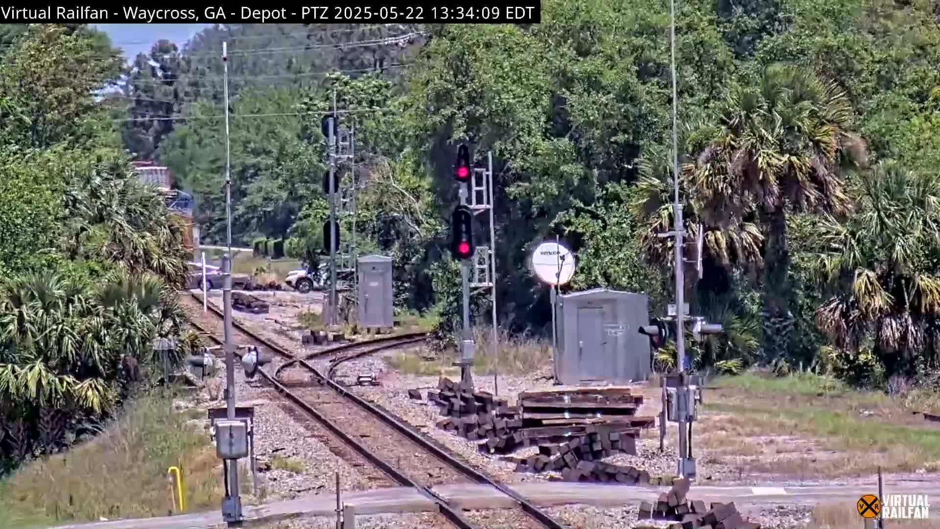 A train is partially visible behind some trees near a railroad crossing with a red signal light; the weather is sunny.