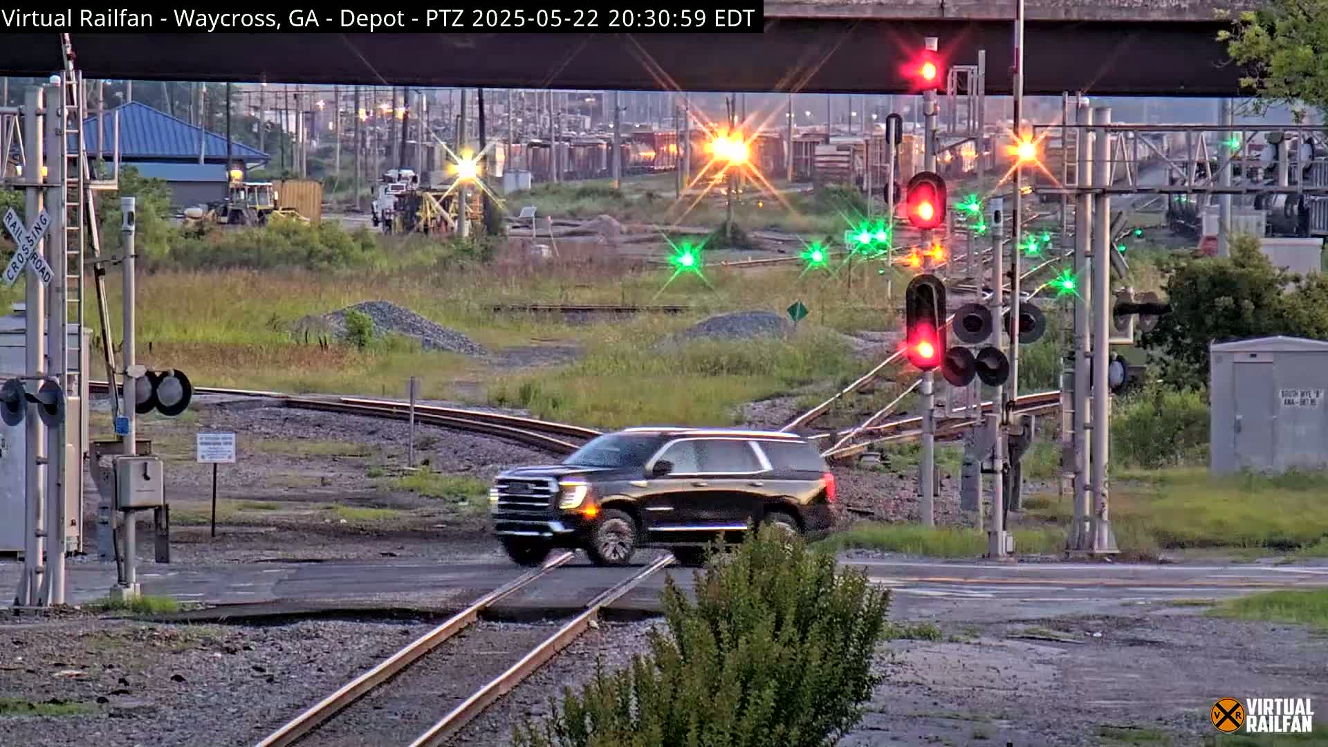 A dark-colored SUV crosses a railroad crossing under a bridge at twilight, with railroad lights flashing and train cars visible in the background.
