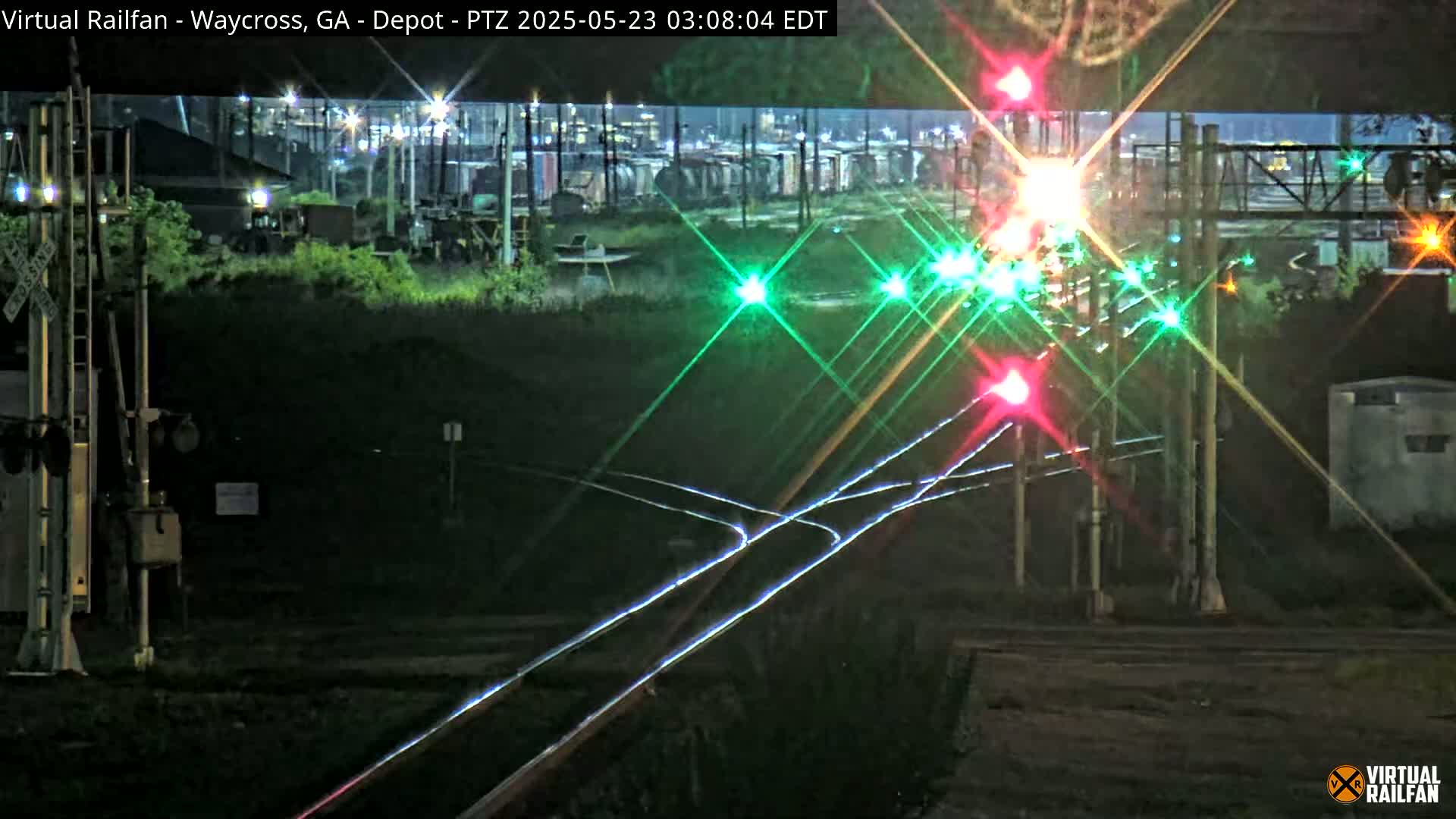 At night, illuminated railroad crossing signals shine brightly near a rail yard with several train cars visible in the distance.