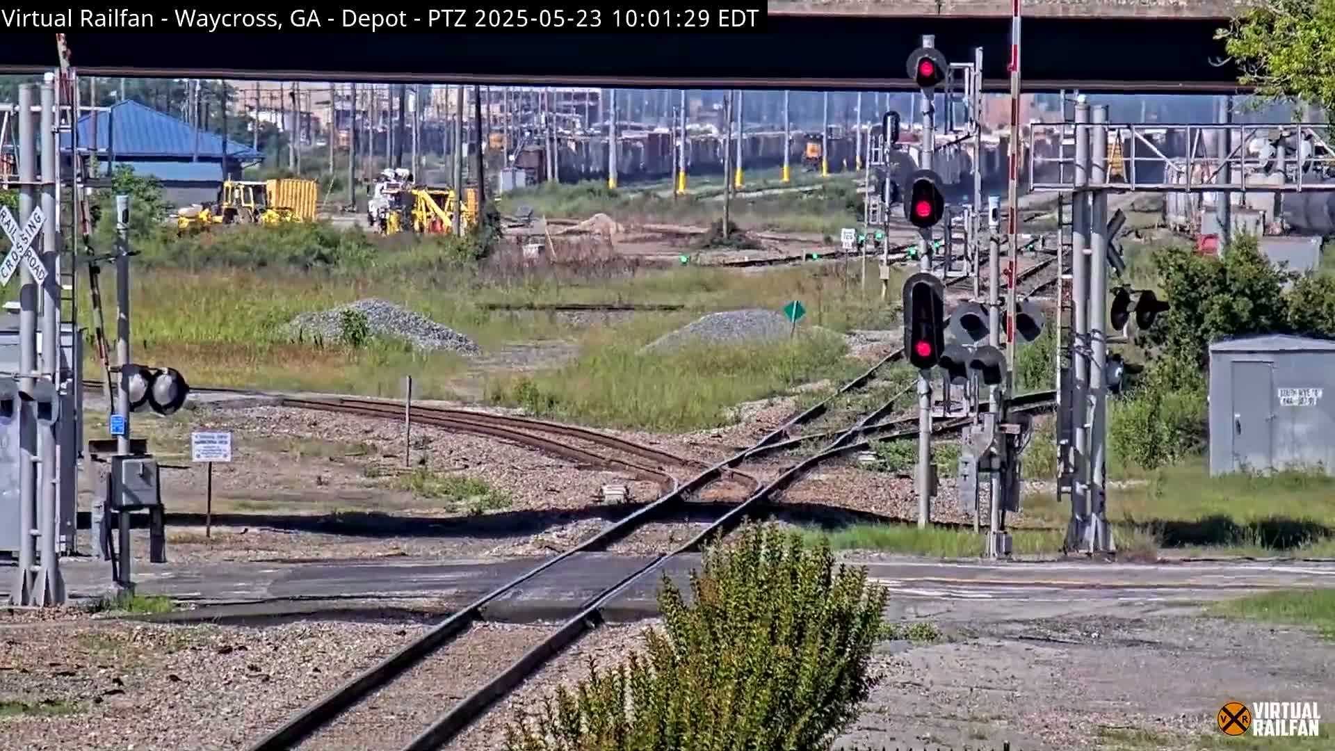 A sunny outdoor scene shows a railroad crossing with several sets of train tracks intersecting, railroad signals, and some construction equipment and buildings in the background.