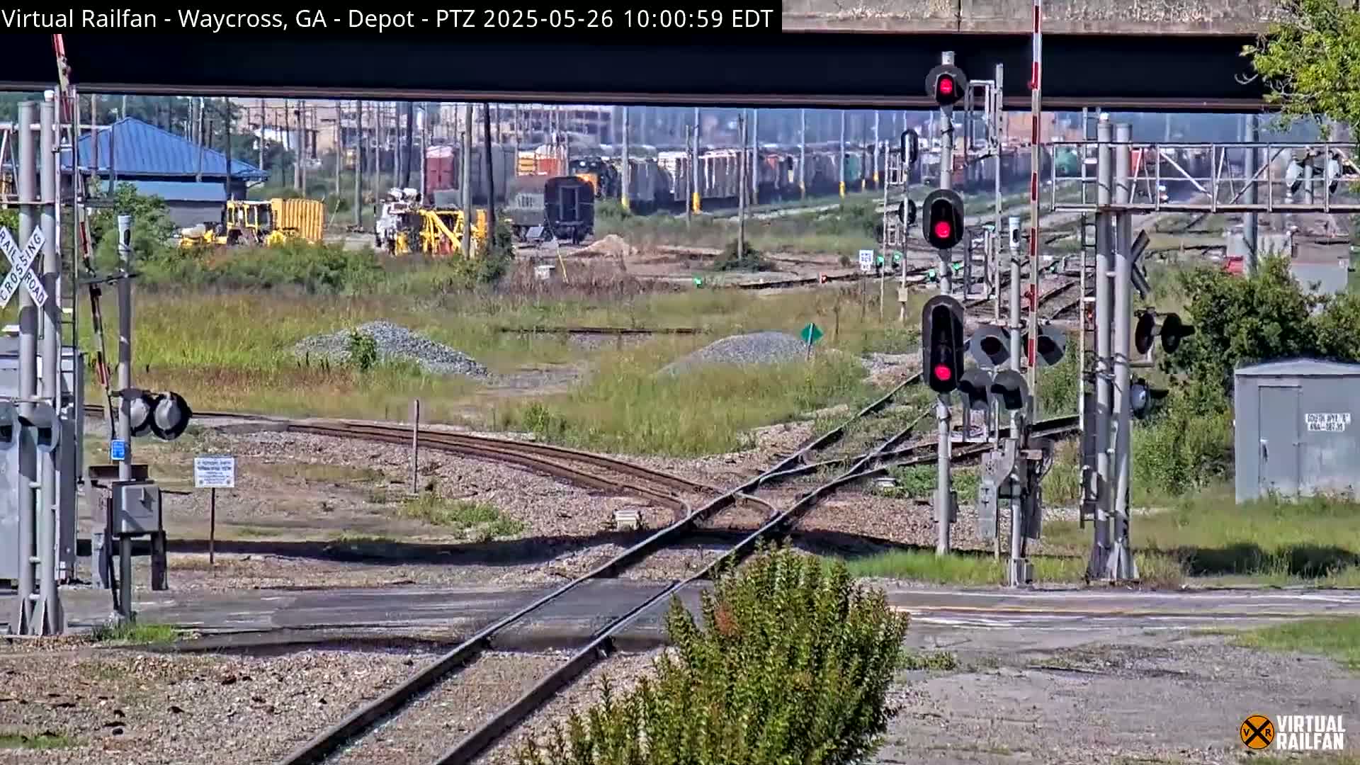 A railroad crossing with multiple sets of tracks, railway signals, and some industrial buildings in the background under a partly sunny sky.