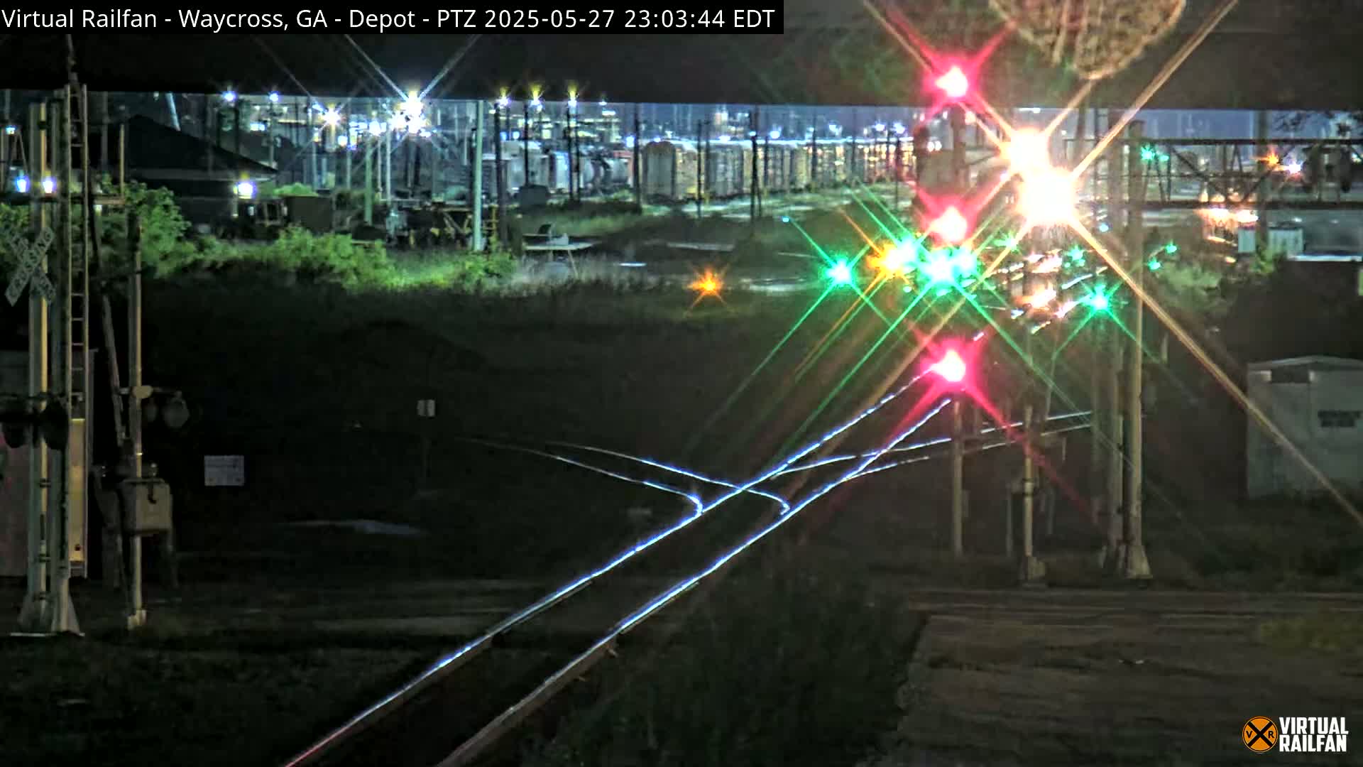 At night, brightly lit railroad crossing signals shine over a dark rail yard with several tracks and some distant buildings.