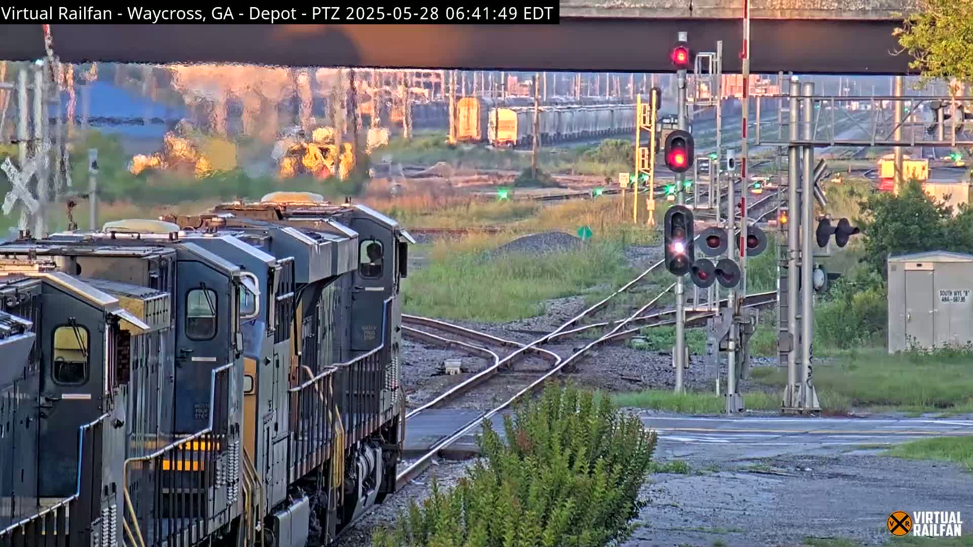 A train of several dark-grey locomotives approaches a railroad crossing under a partly cloudy sky at sunrise.