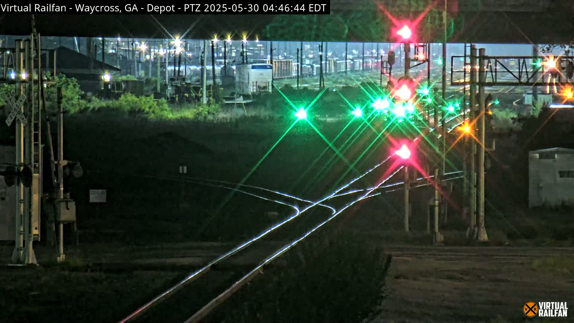 At night, brightly lit railroad crossing signals shine against a dark background, with train cars visible in the distance.