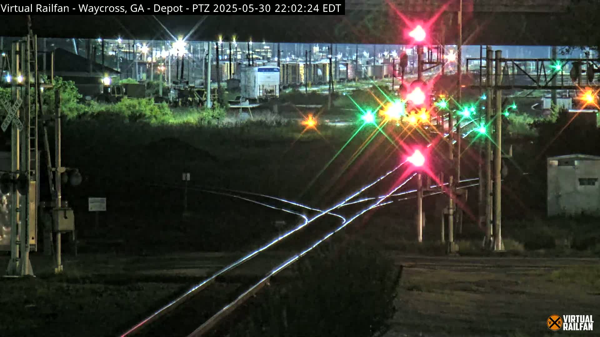 At night, brightly lit railroad crossing signals shine over intersecting train tracks near industrial buildings.