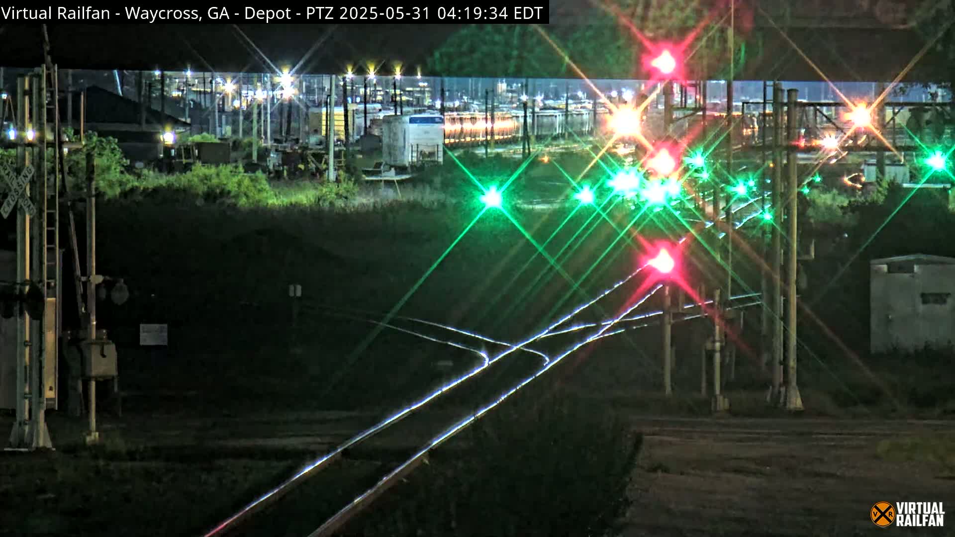At night, a train yard with railroad tracks and flashing red and green signal lights is visible.