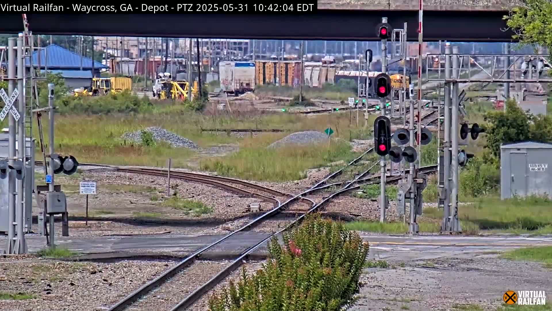 A railroad crossing with several sets of tracks, railway signals showing red, and some industrial buildings in the background on a partly sunny day.