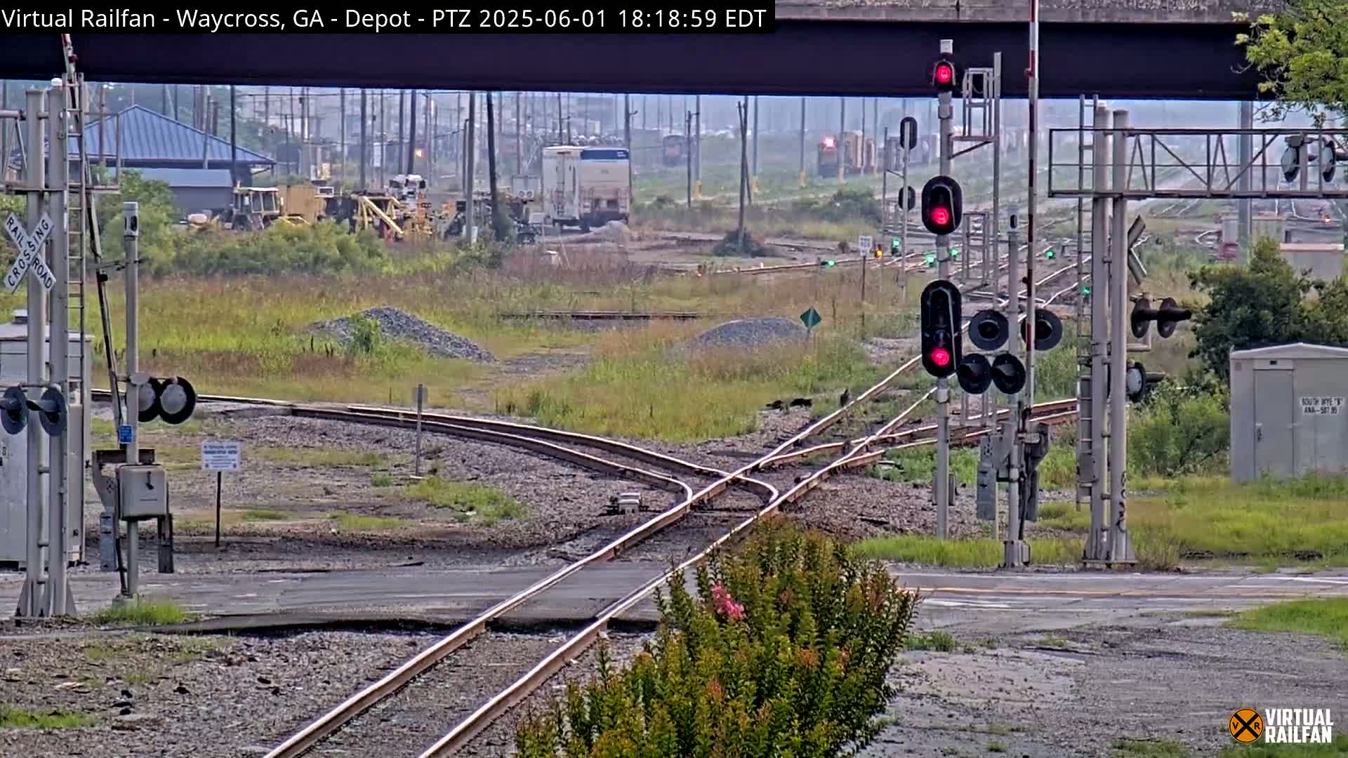 A railroad crossing with several sets of tracks converging, surrounded by overgrown vegetation and a building in the distance, under an overcast sky.