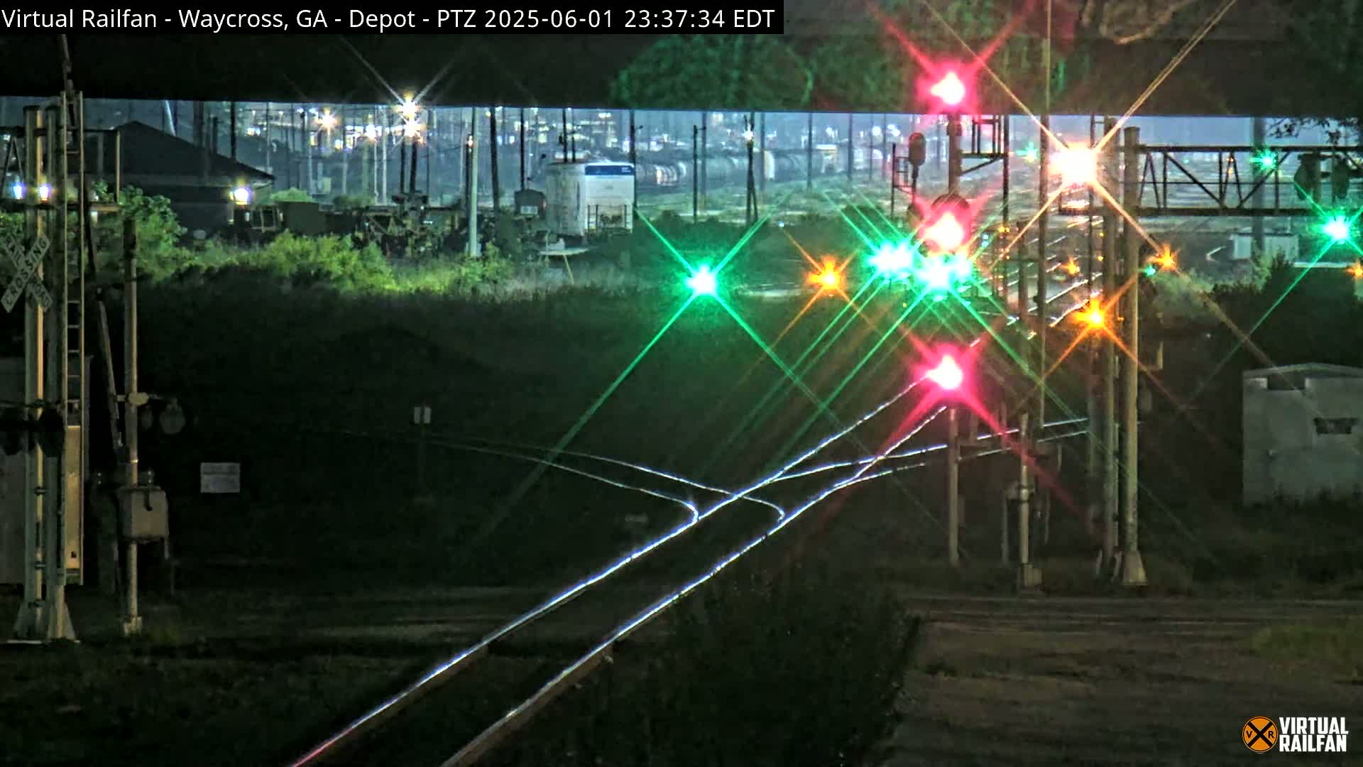 At night, a railroad crossing with multiple sets of illuminated train signals shows a train in the distance under a dark sky.