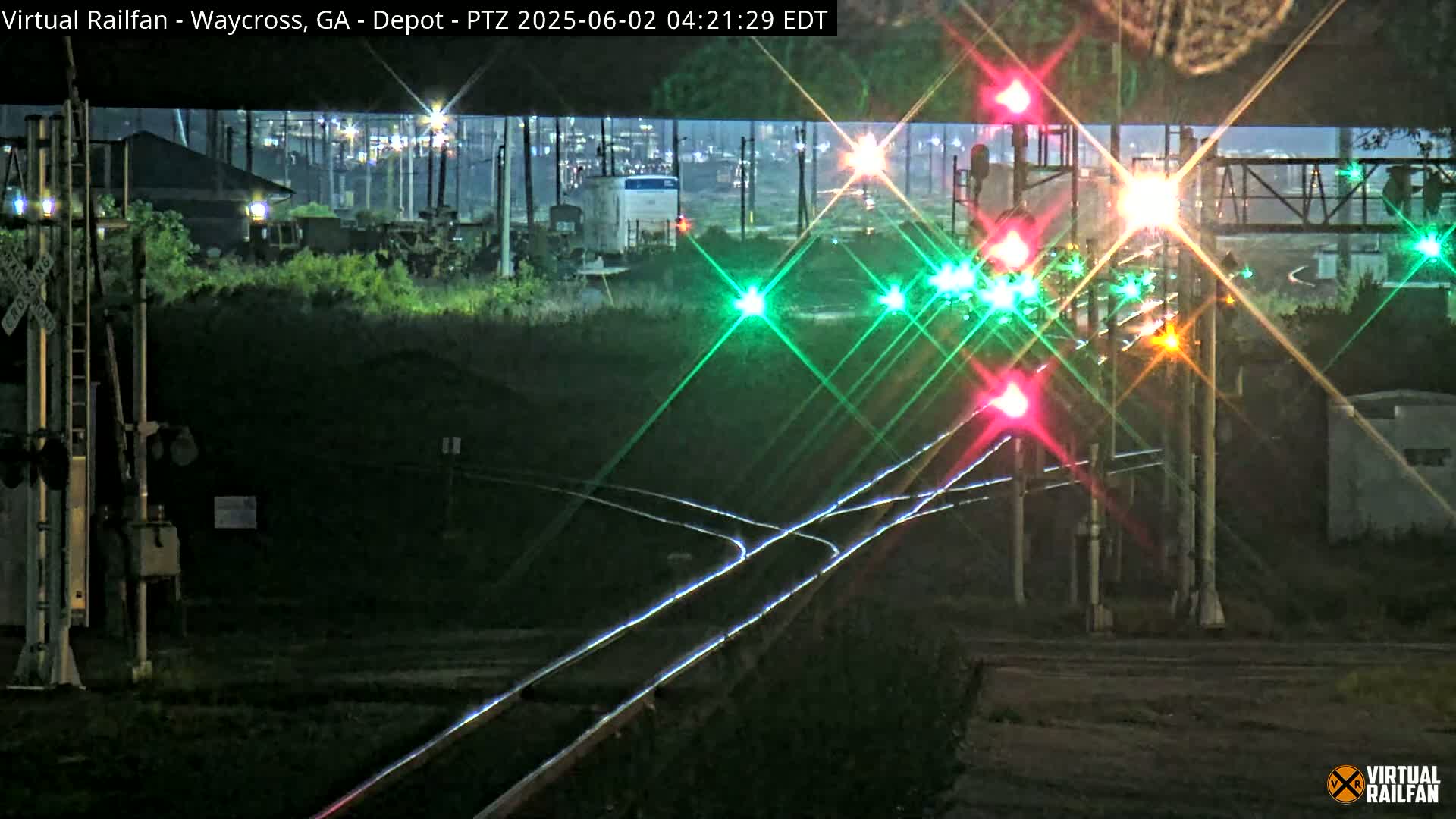 A nighttime view of railroad tracks and signals, with some industrial buildings and vegetation in the background.