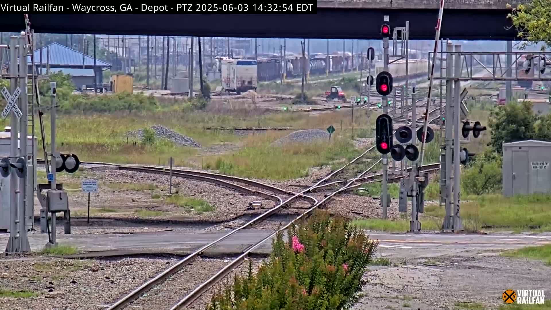 A railway switching yard with multiple train tracks, crossing signals displaying red, and a small utility vehicle is visible on a partly cloudy day.