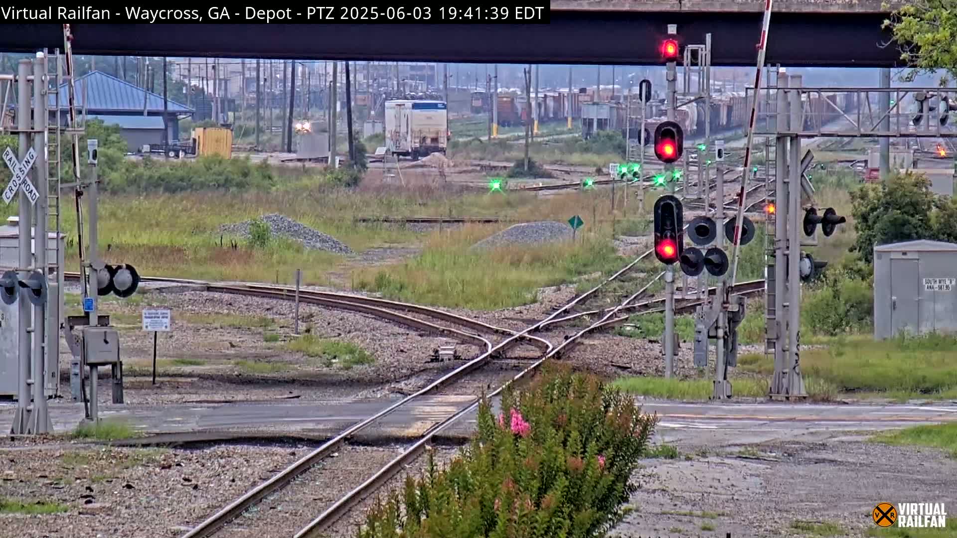 A railroad crossing with several sets of train tracks and traffic signals, under an overcast sky, shows a boxcar in the distance.