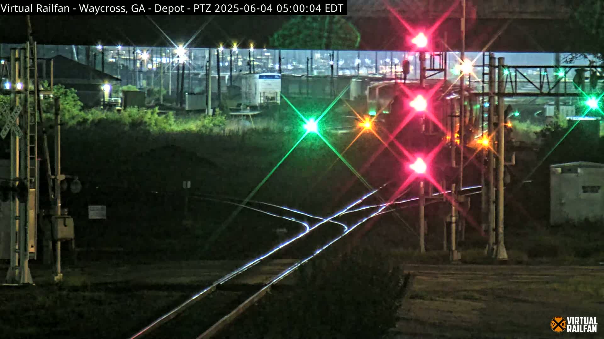 A nighttime view of a railroad crossing with illuminated red and green signal lights, showing train tracks and nearby industrial buildings under a dark sky.
